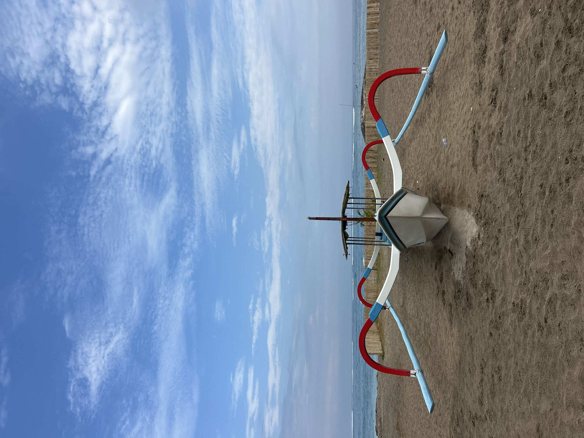 A front-facing, medium shot captures a traditional Balinese outrigger boat resting on the sandy shore of Kuta Beach, Indonesia. The boat is predominantly white with striking red and blue outriggers extending outwards. The ocean, with a calm horizon and faint waves, serves as the backdrop, while a bamboo fence lines the edge of the beach. The sky above is a vibrant blue, dotted with wispy white clouds. The scene is devoid of any people.