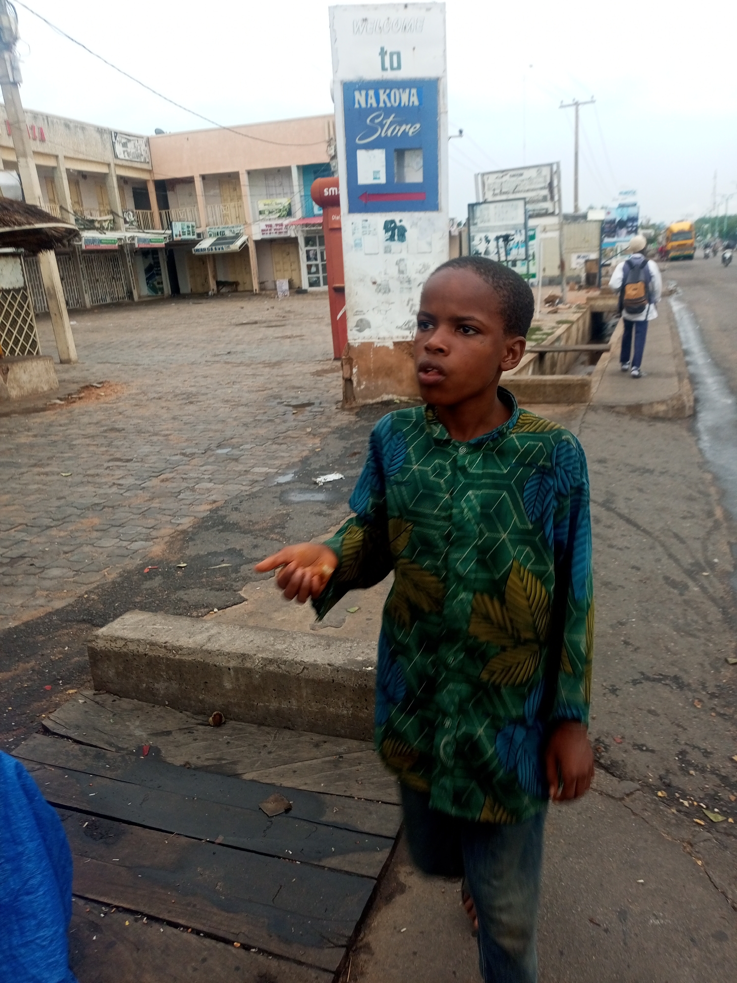 A young African boy stands near a storm drain in Gombe, Nigeria. He wears a green and blue patterned shirt and jeans. A sign reading "NAKOWA Store" is visible in the background. A person with a backpack walks along the road in the distance. Shops line the street. The sky is overcast.