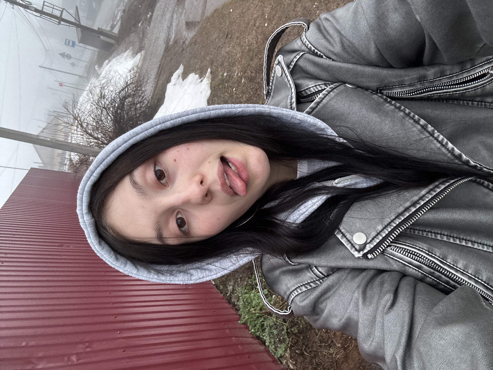 A young woman with dark hair and brown eyes is taking a selfie. She is wearing a grey hoodie and a metallic silver jacket. Her tongue is slightly extended. The background shows a hazy, overcast environment, suggesting fog or mist. In the distance, a street sign and poles are visible, and there is some residual snow on the ground. To the left, a red corrugated metal fence is visible. The surroundings suggest a location in a city, possibly Фоки, Russia, given the query.