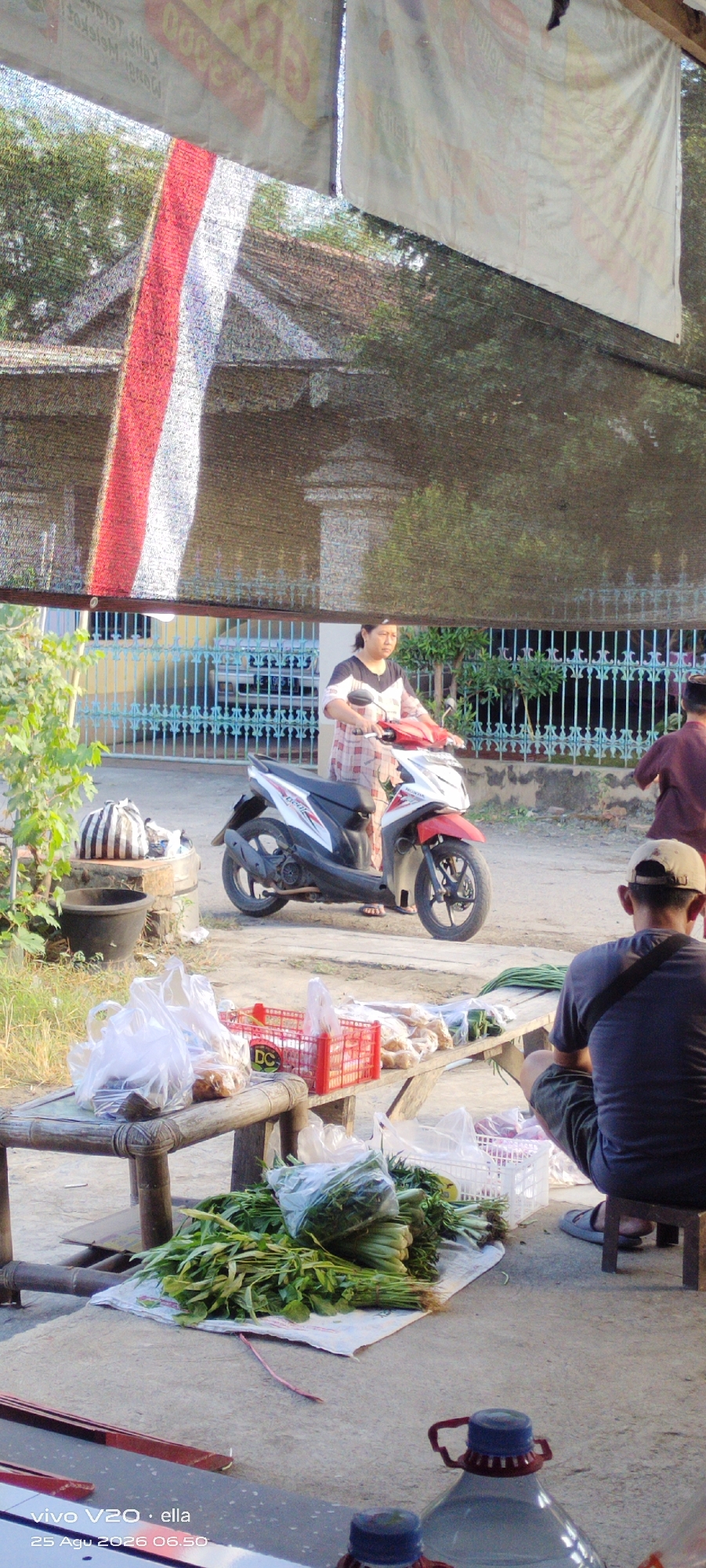 A medium shot shows an outdoor scene in Simo, Indonesia, with a woman on a scooter and a seated man. The woman, wearing a plaid shirt and dark pants, is positioned centrally, operating a red and white scooter. In the foreground, a man wearing a dark t-shirt, shorts, and a cap is squatting, facing away from the camera, looking at produce displayed on a wooden bench. The bench holds several plastic bags filled with vegetables, red baskets, and packaged food items. Behind the woman, a blue metal fence runs along a sidewalk, with a building and trees visible beyond. Overhead, a black mesh shade covers part of the scene, with a red and white striped banner visible at the top left. The ground is concrete and dirt. The timestamp "25 Agu 2026 06.50" is present in the lower left corner.