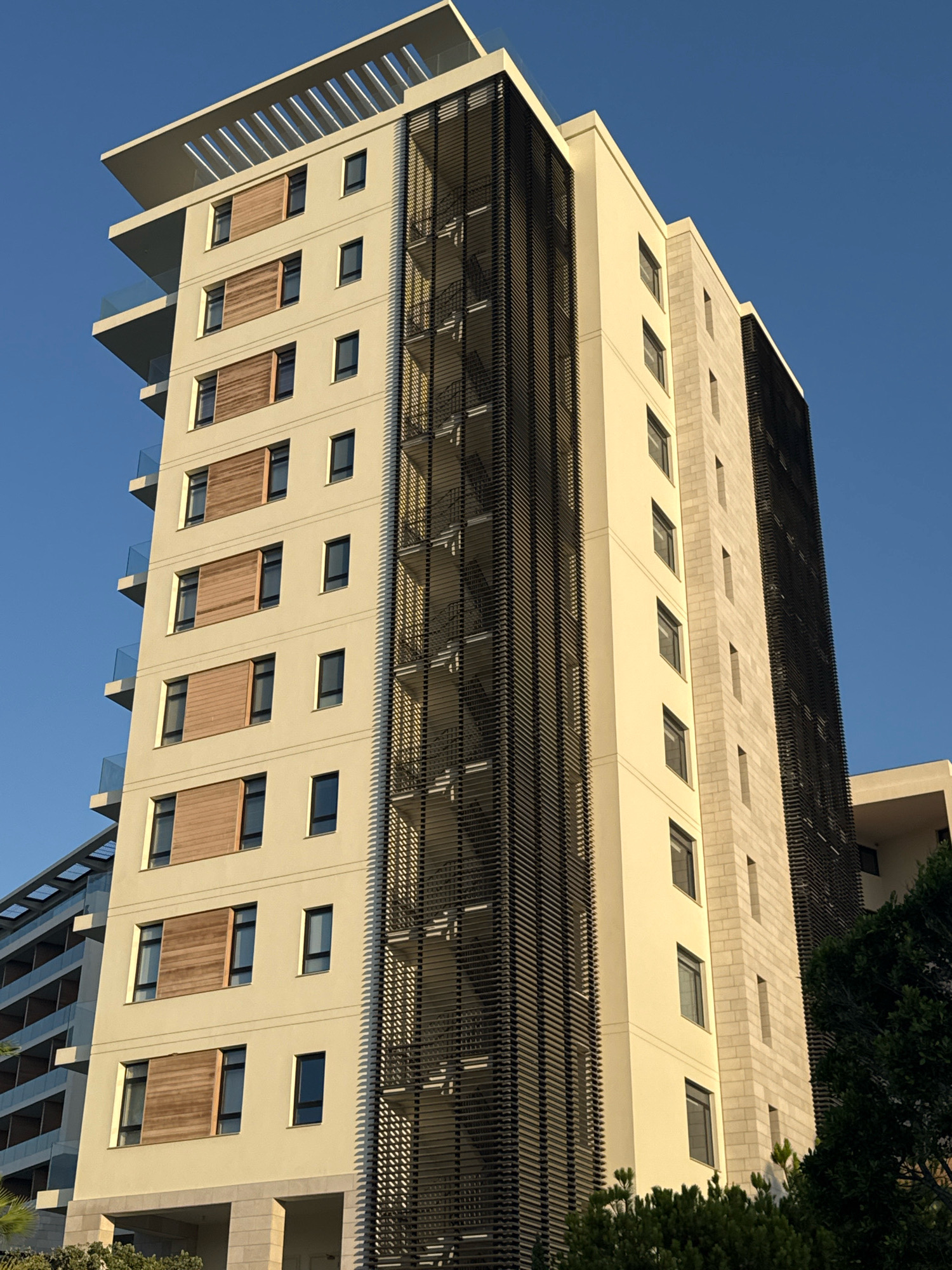 A low-angle, medium shot captures a modern, multi-story apartment building against a clear, bright blue sky. The building features a cream-colored facade with multiple balconies, each equipped with clear glass railings. Several windows are visible, some with wooden shutters. A vertical structure with thin, dark metal slats runs along a portion of the building's side. To the left, another building with a similar facade and balconies is partially visible. The foreground shows lush green trees and foliage. The image is set in Agios Tychona Community, Cyprus.