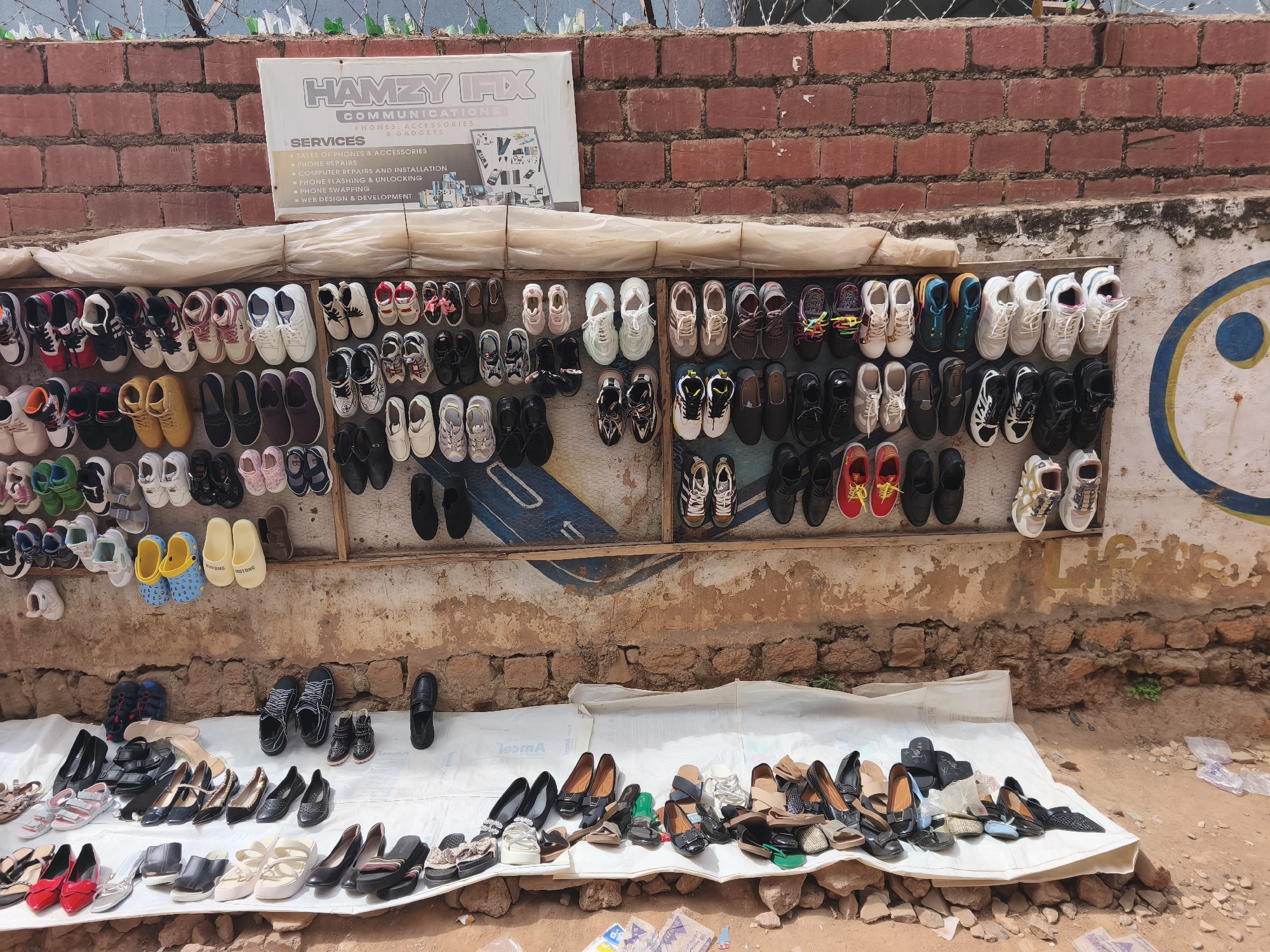 An outdoor market stall in Jos, Nigeria, displays a wide array of footwear against a backdrop of brick and painted walls. Numerous pairs of sneakers, including athletic and casual styles in various colors like white, black, red, yellow, and multi-colored laces, are hung along wooden rails. Some yellow clogs are visible, with "HUTONG" printed on them. The shoes are attached to a weathered, white-painted wall featuring a faded blue and yellow circular motif. Above this, a red brick wall topped with barbed wire and broken glass suggests a boundary.
On the ground, a white tarp is spread over dirt and rocks, showcasing a diverse collection of women's shoes, including elegant black flats, patterned sandals, and slip-ons. The arrangement suggests a bustling market environment.
A prominent white sign, "HAMZY IFX COMMUNICATIONS," is affixed to the brick wall, advertising "PHONES, ACCESSORIES & GADGETS." Below this, a list of "SERVICES" includes "SALES OF PHONES & ACCESSORIES," "PHONE REPAIRS," "COMPUTER REPAIRS AND INSTALLATION," "PHONE FLASHING & UNLOCKING," "PHONE SWAPPING," and "WEB DESIGN & DEVELOPMENT." The scene is brightly illuminated by daylight, indicating a sunny outdoor setting. A faint "2009" is visible on the upper left section of the painted wall.