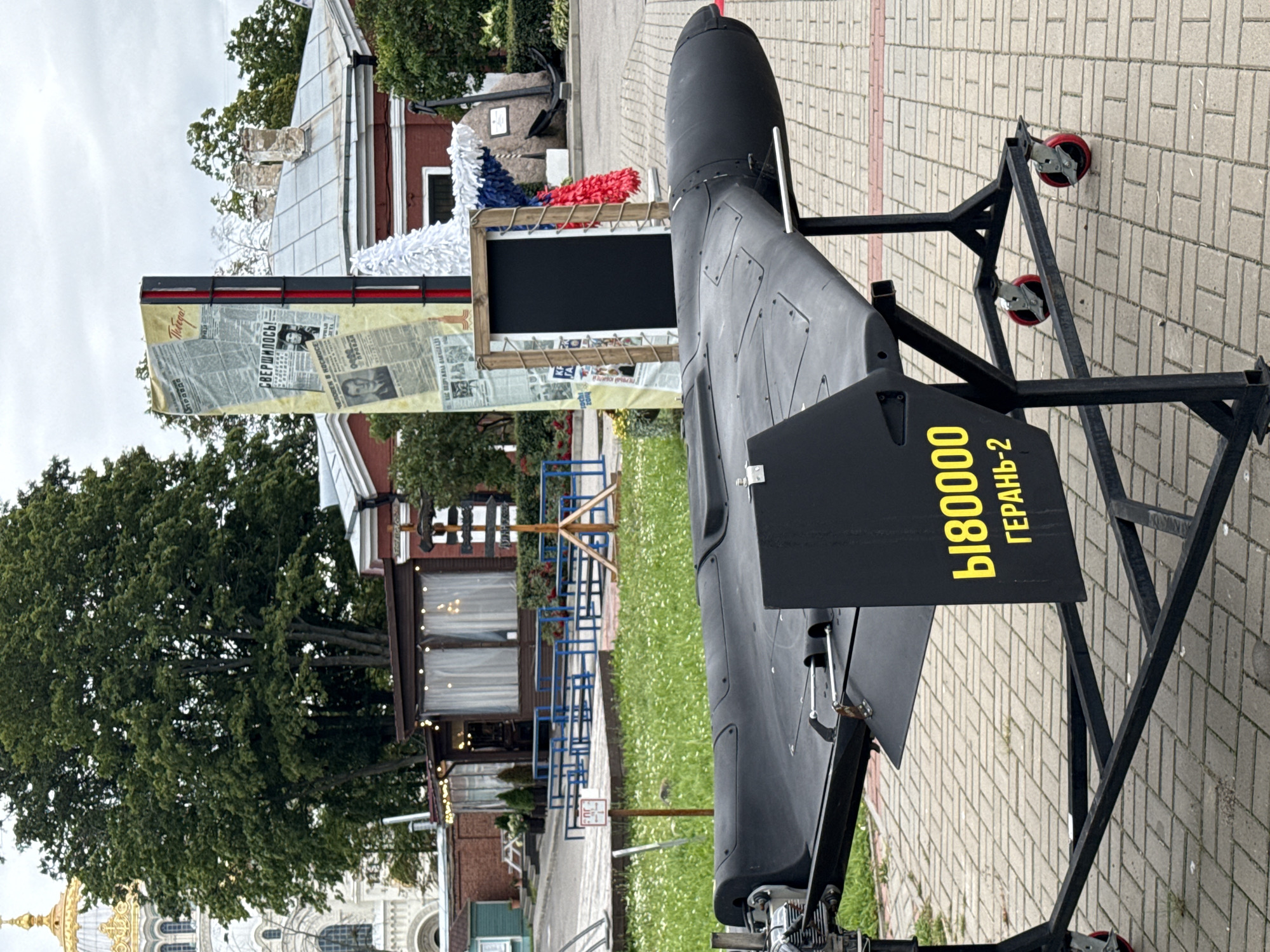 The image depicts a drone on display in an outdoor urban setting. The drone is predominantly black with yellow lettering on its tail fin that reads "Ы180000 ГЕРАНЬ-2". The drone is mounted on a black metal stand with wheels. In the background, there is a building with a golden dome, likely a church or cathedral, and other structures. A large tree with green foliage stands prominently. A tall display board with what appears to be newspaper clippings and images is also visible. The ground is paved with brick-like patterns and a grassy area with scattered white flowers. The scene appears to be in the city of Kronstadt, Russia, as indicated by the architectural style and the context of the drone, which has been associated with military operations. The weather seems overcast. There are no people in the immediate foreground or clearly interacting with the drone.