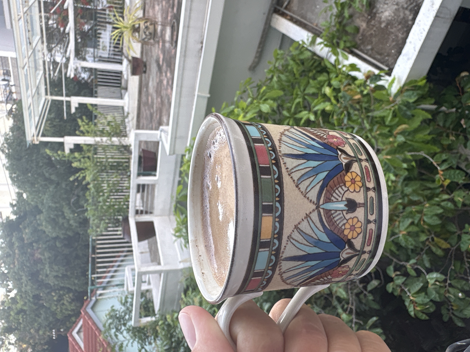 An outdoor scene at what appears to be a rooftop or balcony in Nha Trang Ward, Vietnam, is captured. A person's hand is holding a ceramic mug filled with a frothy, light brown beverage, likely coffee, in the foreground. The mug itself is decorated with an intricate pattern featuring stylized blue and green foliage elements, yellow and red floral motifs, and a geometric band in various colors along its rim. In the background, lush green foliage and trees dominate the view. Further back, architectural elements suggest a multi-level structure, possibly a building with a white pergola-like roof and white railing. Several potted plants are visible on the upper level, and the ground is paved with reddish-brown tiles. The overall lighting suggests it is daytime, possibly late afternoon or early morning due to the soft light.