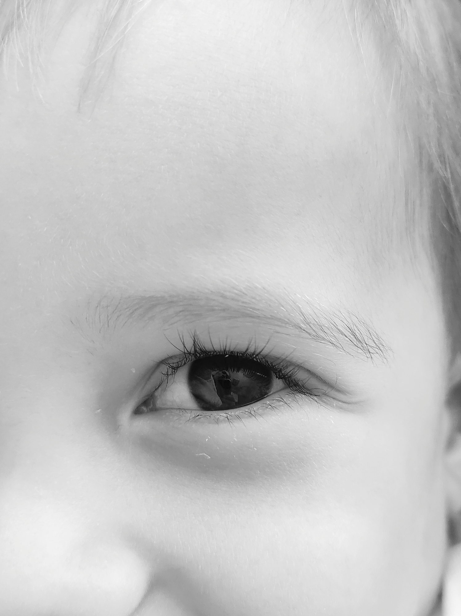 A close-up, black and white photograph shows the left eye of a young child, with a portion of their forehead and cheek visible. The child has fair skin and light-colored hair, some of which falls across their forehead. The eye itself is dark, with long eyelashes. The image focuses on the texture of the skin and the details of the eye, creating an intimate and detailed portrait. The background is largely out of focus, emphasizing the subject's eye.