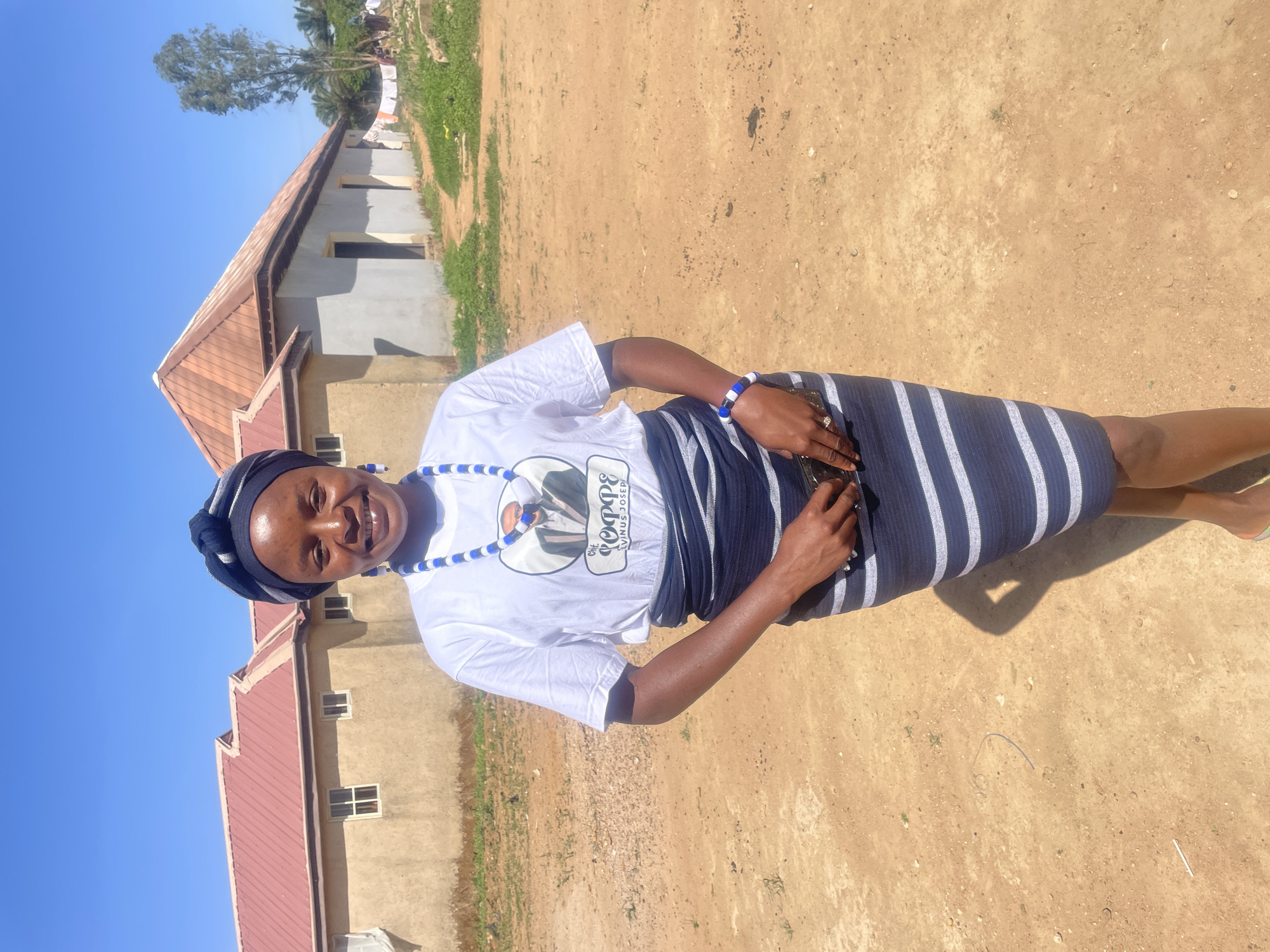 A woman is standing in an outdoor setting, likely Durimi, Nigeria. She is wearing a white T-shirt with an image of someone printed on it, a blue and white striped skirt, a blue and white beaded necklace, a blue and white bracelet, and a head wrap with the same color scheme. She is holding an object in her right hand. Behind her, there is a building with a reddish-brown roof and another building in the distance, along with a tree. The ground is a reddish-brown color. The sky is a clear, bright blue, suggesting a sunny day.
