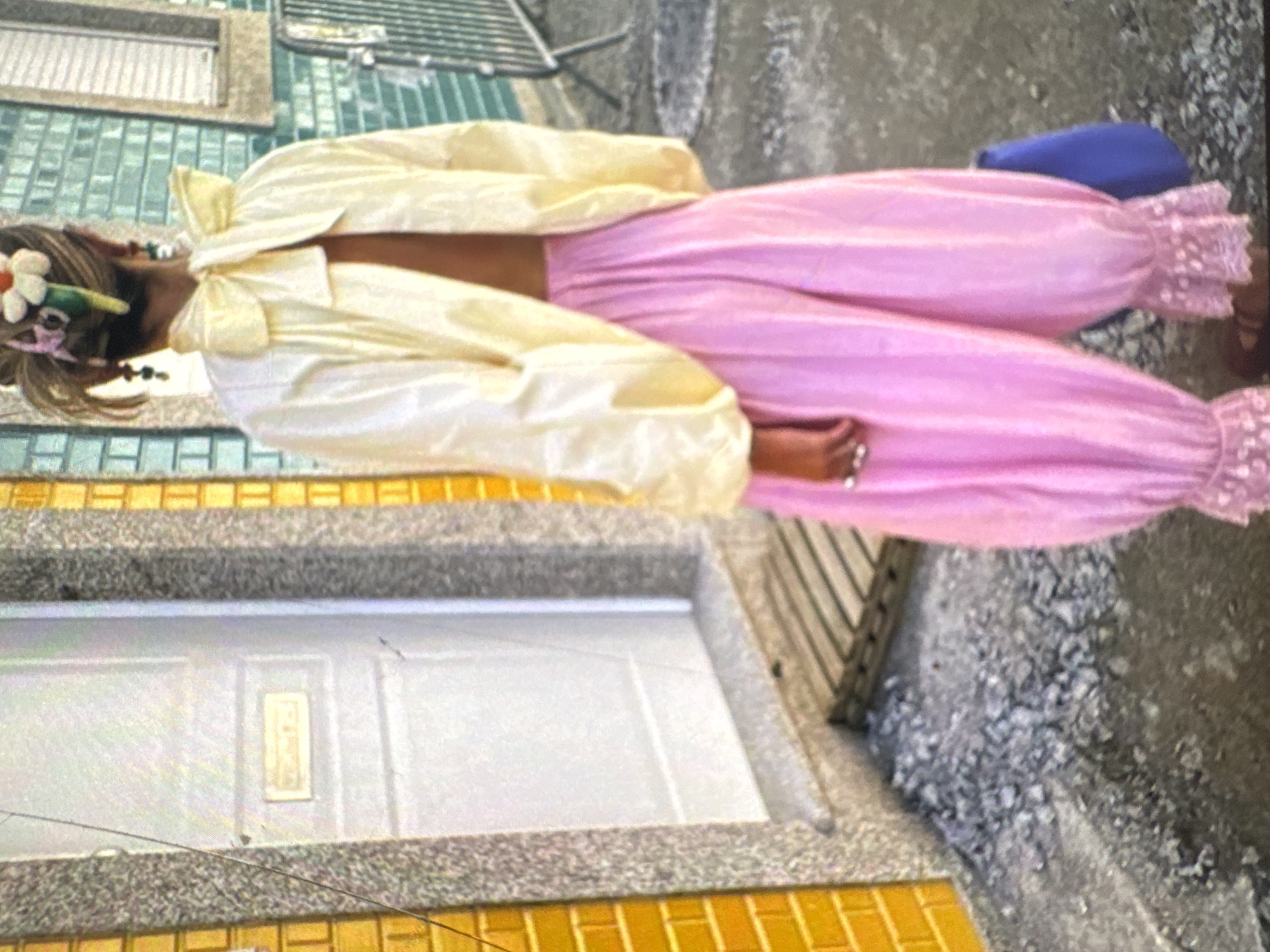 The image depicts a woman from behind, standing outdoors in what appears to be an urban environment. She is wearing a cream-colored, open-back top with a large bow at the neck and voluminous pink satin pants that are gathered at the ankles with lace trim. She has a flower accessory in her hair. The background includes a white door with a metal mail slot, yellow brickwork, and a green tiled wall. The ground is covered in gravel and what appears to be wooden planks. A blue bag is visible near her feet. The setting does not provide any specific visual cues to confirm a location in Protasovo, Russia.