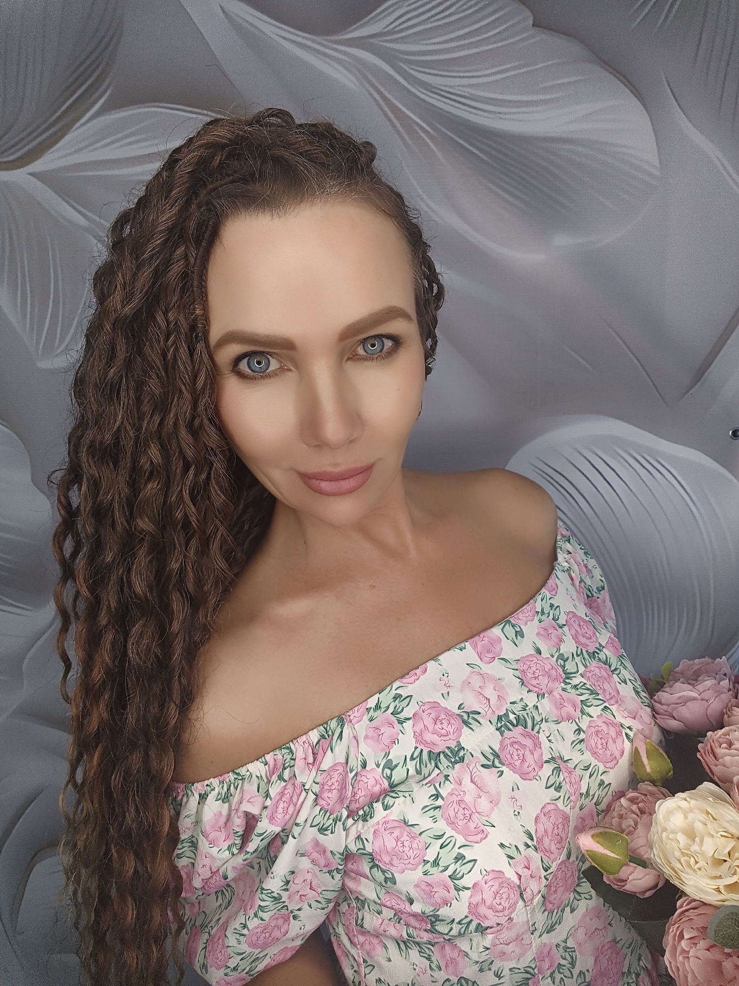 A woman with long, curly brown hair and blue eyes is looking directly at the camera. She is wearing an off-the-shoulder white dress with a pink floral pattern. She is holding a bouquet of pink and cream-colored flowers. The background is a light gray with a subtle floral pattern. The image appears to be a portrait, possibly taken indoors. The woman's expression is serene. The lighting suggests it may be daytime, but there are no strong indications of the time of day or weather. The location is not specified. There is no visible text in the image.