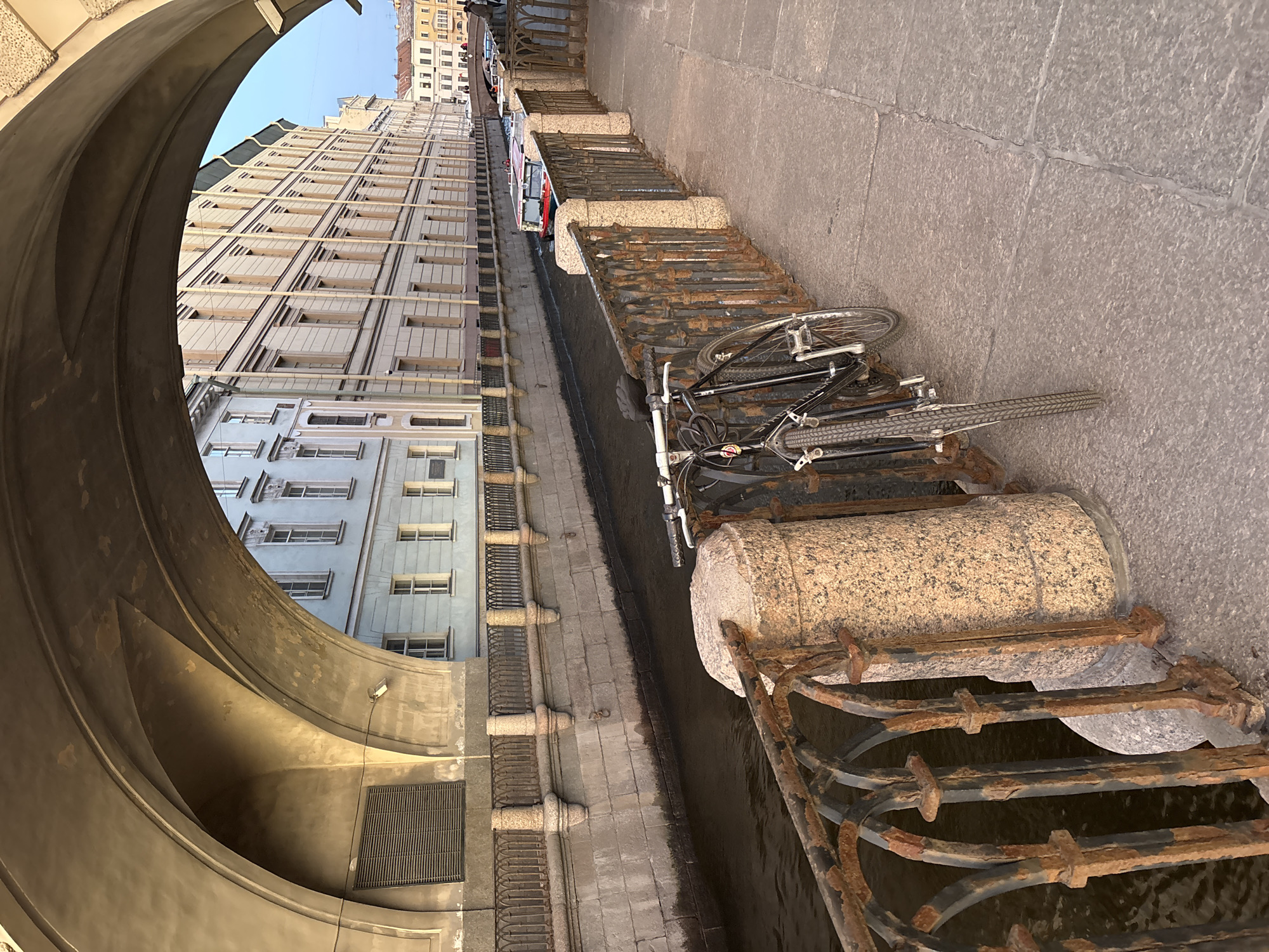 The image captures a scene in Saint Petersburg, Russia. The view is taken from under an arch, framing a perspective along a canal. Parallel to the canal, a row of buildings rises, showcasing a variation of architectural styles and window arrangements. A rusty metal railing with stone pillars runs along the canal's edge. A bicycle lies horizontally across the railing. The canal water is dark. The image shows the interplay of architectural elements and urban infrastructure in Saint Petersburg.