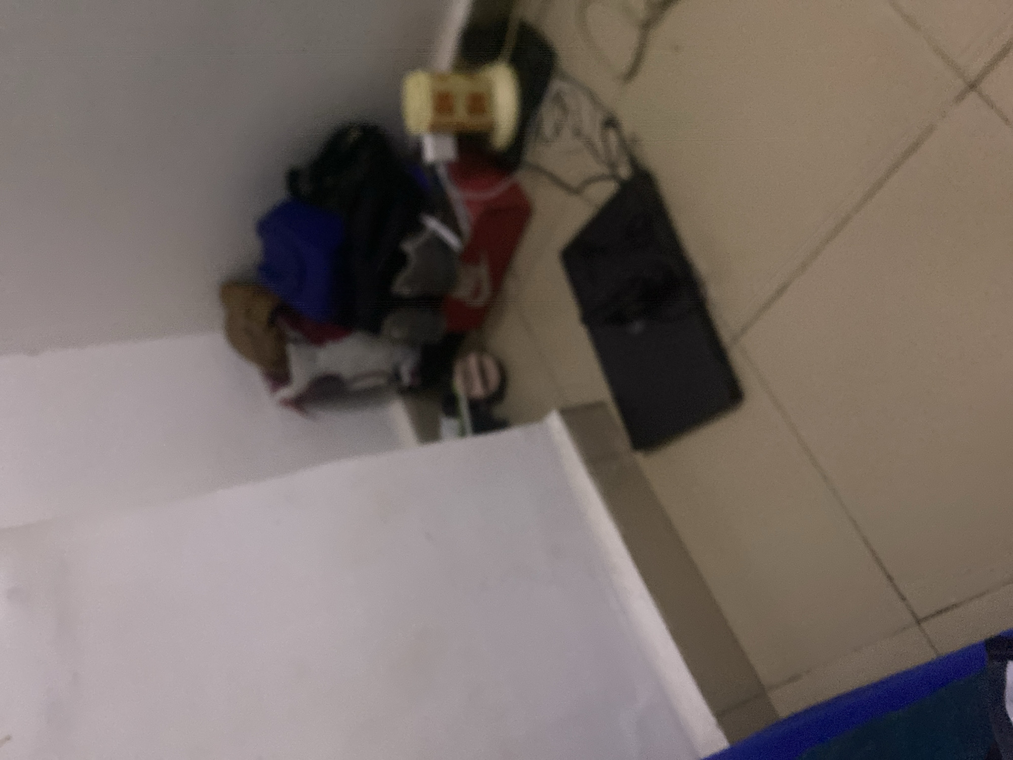 The image shows a cluttered corner of a room. A white wall meets a tiled floor at a baseboard. In the corner, a pile of bags and clothing is visible, including a blue backpack, a tan item, and a red bag. Next to this pile, a small, cylindrical object with an orange top sits on a black mat. A black, rectangular electronic device is on the floor in the foreground. To the left, a portion of a blue and black object, possibly furniture or clothing, is visible. The lighting is dim, and the image is slightly out of focus. The specific location in the city of Howayi, Nigeria, cannot be determined from the provided image.