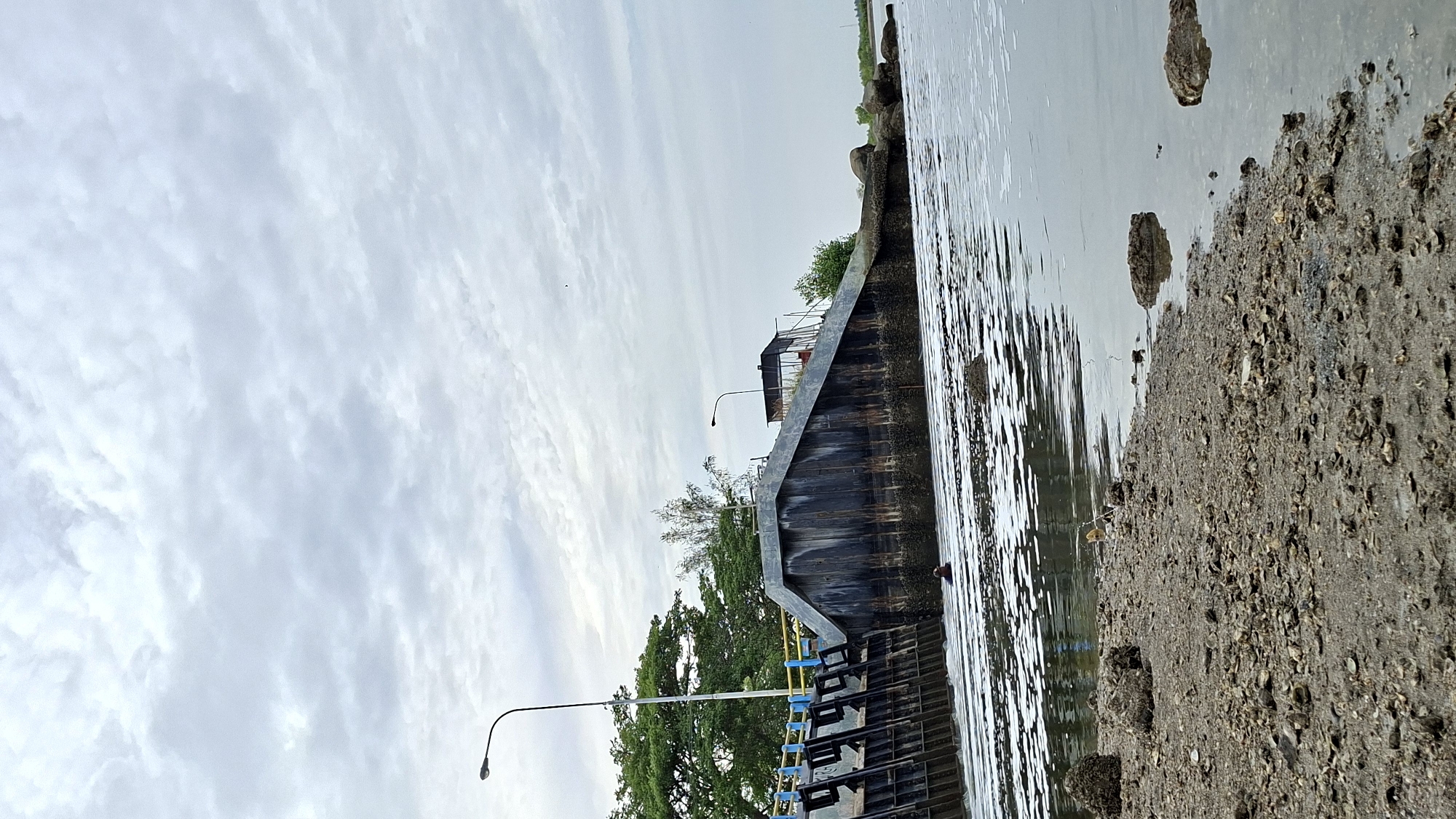 This image captures a view of the Krueng Geukueh Dam in Lhokseumawe, Indonesia. The dam, a significant infrastructure in the city, is a prominent feature with its metal gates and concrete structure. The water is disturbed, creating white foam along the dam's edge, suggesting water flow. In the distance, a person is partially visible in the water, near the dam. The foreground shows a muddy bank covered with shells and rocks, indicating a natural shoreline. The sky is overcast with a dense cloud cover, and trees are visible in the background on the left side of the dam. Two lampposts are also present, one on the left and another on the right side of the dam. The overall scene depicts a functional water control structure within a natural, albeit somewhat industrial, environment.
