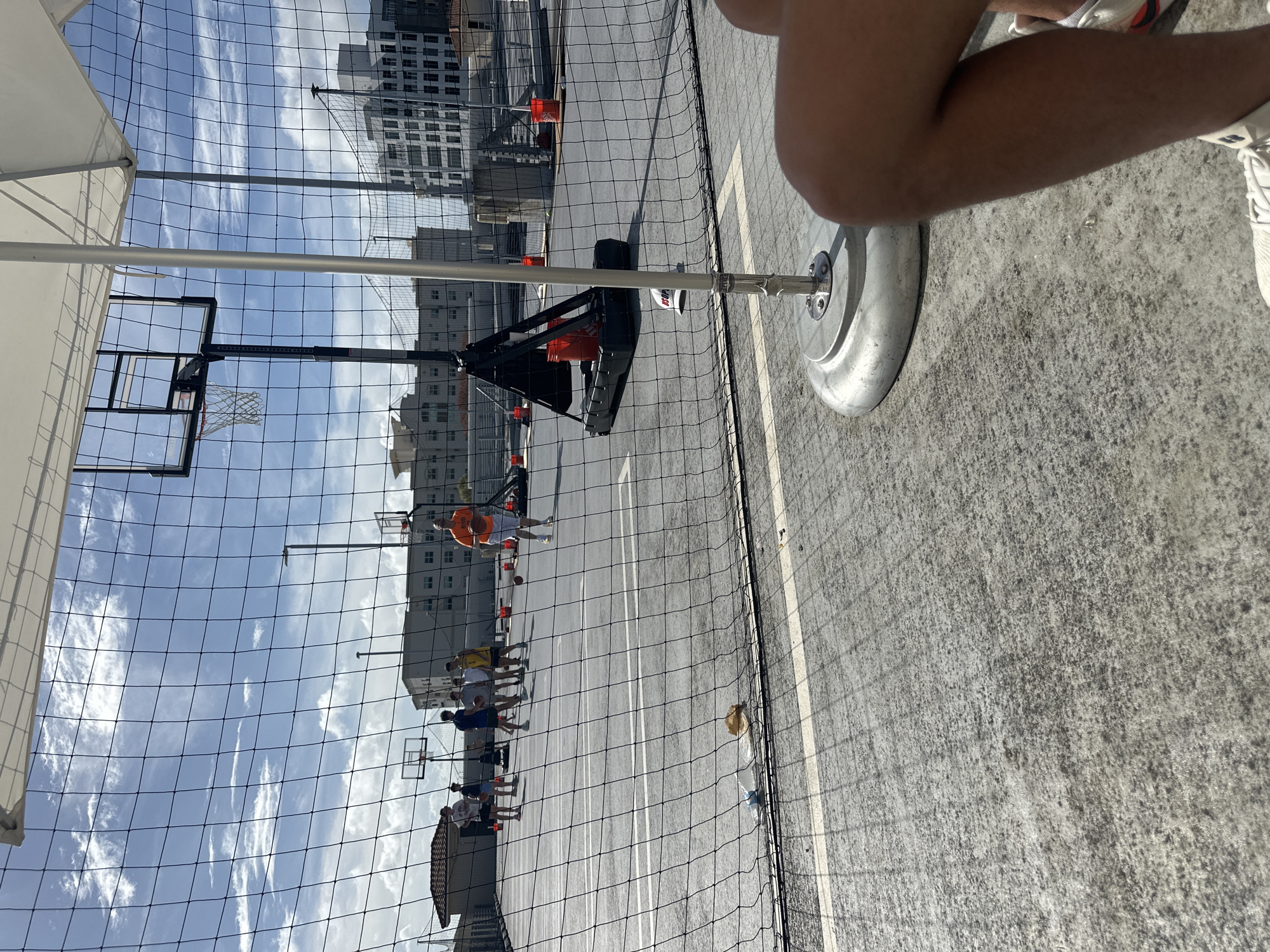 The image captures a scene on an outdoor basketball court, possibly within the city of Aventura, United States of America, given the urban architecture in the background. A group of people, appearing to be playing or practicing basketball, are visible on the court. One individual in an orange jersey is holding a basketball. The basketball court is enclosed by a net. A portable basketball hoop is set up, and the base of the hoop is prominently featured in the foreground along with a portion of an individual's leg and shoes.