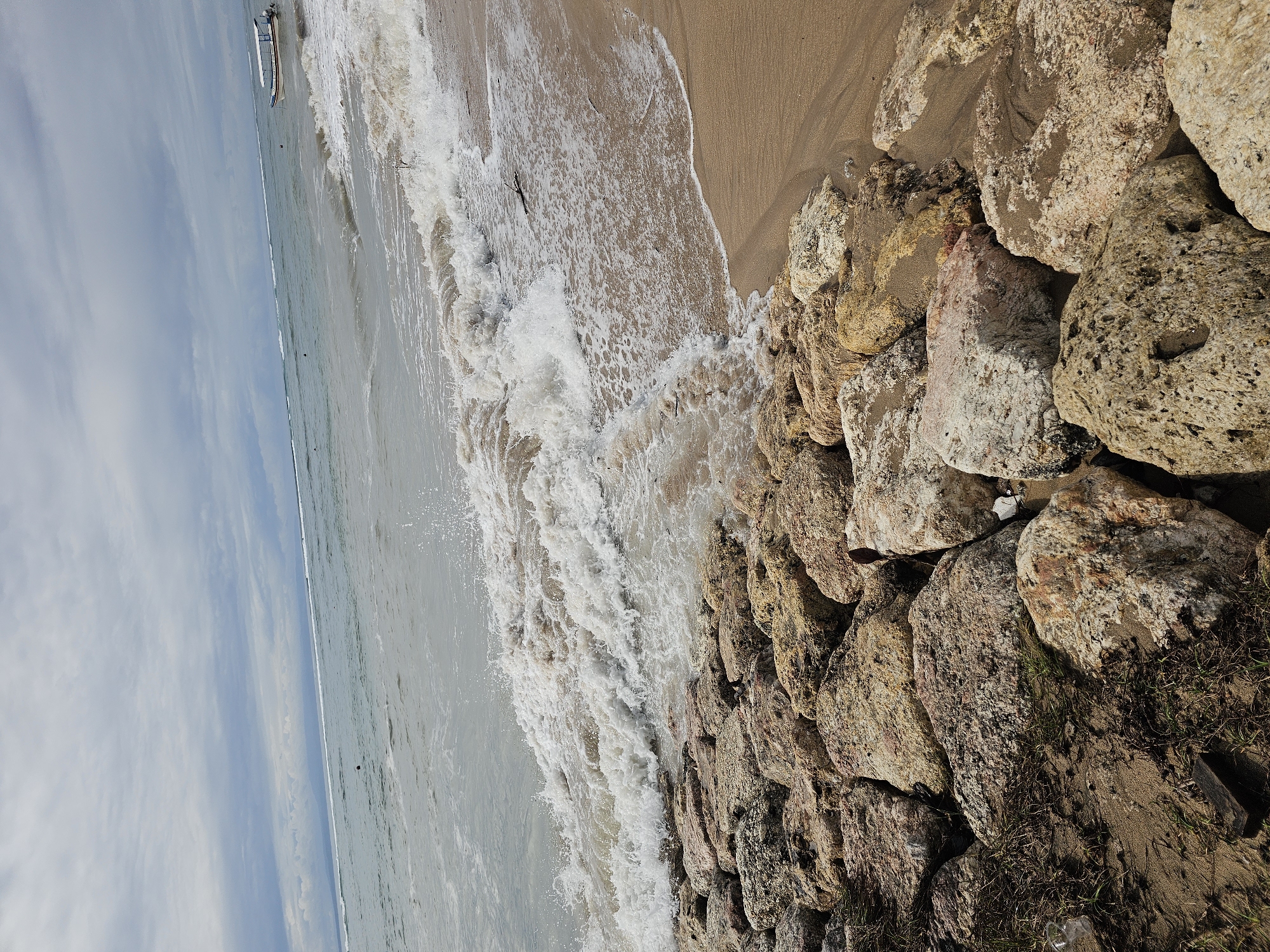 The image shows a beach scene in Denpasar, Indonesia. Waves are crashing against a shoreline reinforced with large rocks. A sandy beach is adjacent to the rocks. In the distance, a boat is visible on the water under a cloudy sky.