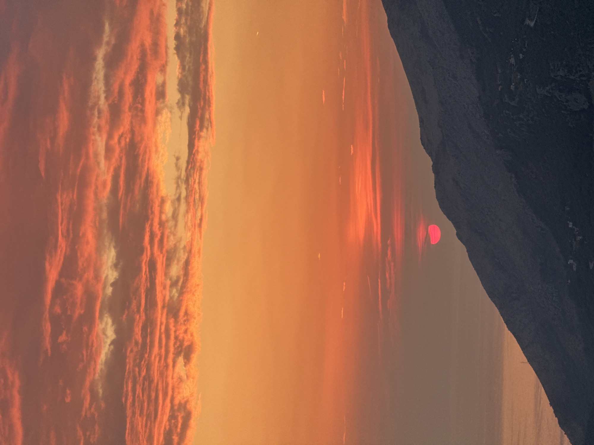 A vibrant sunset paints the sky in hues of orange and red, casting a warm glow over a mountainous landscape. The sun, a bright red disc, is partially obscured by a silhouetted mountain range on the right side of the frame. Wispy clouds stretch across the sky, catching the last rays of sunlight. The ocean is visible in the lower left corner, reflecting the colorful sky. The scene appears to be a natural landscape, possibly a coastal area with mountains. The image does not depict any people or specific objects that would indicate a particular city or urban environment.