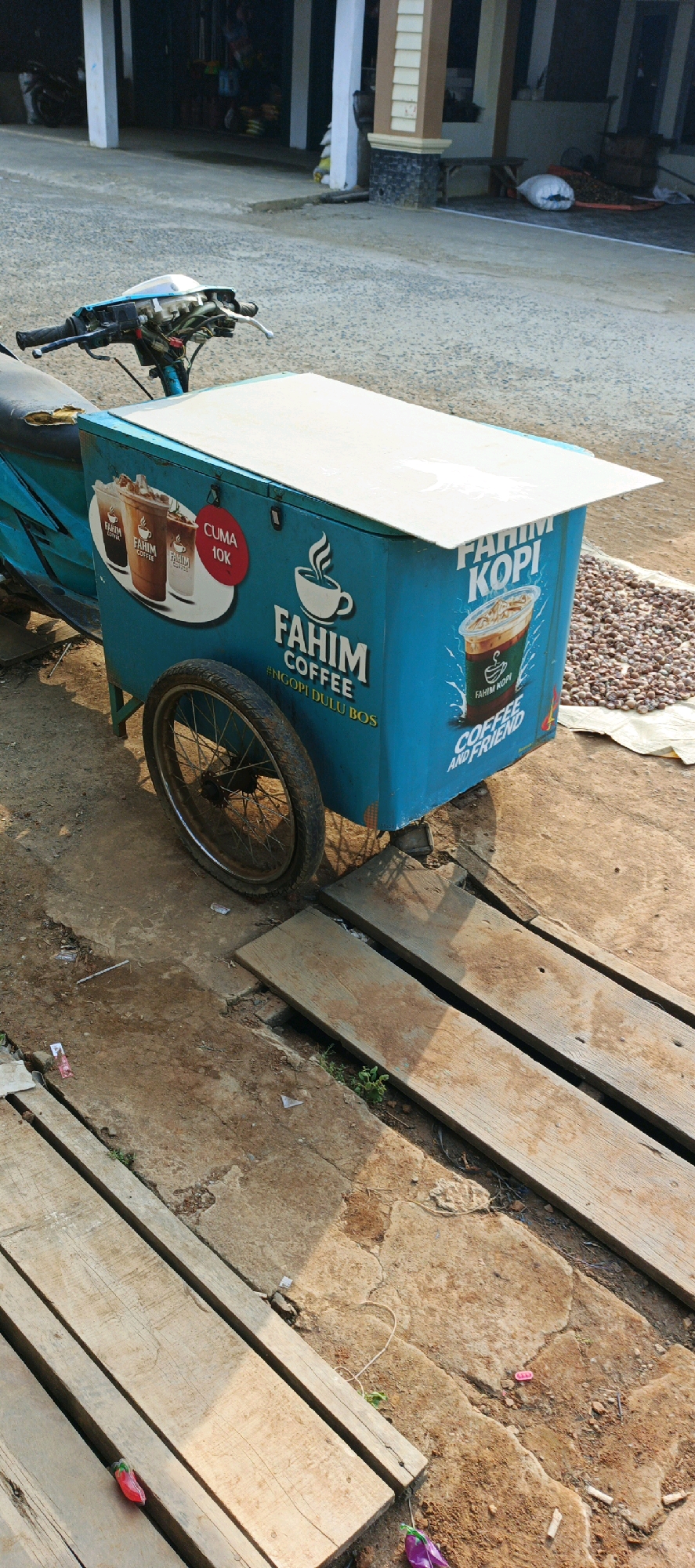 The image depicts a mobile coffee cart, likely a "Fahim Kopi" vendor, parked on a concrete surface in Simbur Naik, Indonesia. The cart, painted bright blue, is attached to the rear of a motorcycle, which is partially visible. The cart features the "Fahim Kopi" logo prominently displayed, along with images of coffee drinks and the tagline "#NGOPI DULU BOS" (roughly translated to "Have coffee first, boss"). A red circular sticker indicating "CUMA 10K" (meaning "only 10,000") is also visible, suggesting a price point.
The immediate surroundings show a worn, dusty concrete ground with wooden planks laid across it in places, possibly to create a more stable or level walking surface. To the right of the cart, a pile of small, dried, brown objects, possibly coffee beans or similar produce, is spread out on a piece of white cloth. In the background, a building with white pillars and a darker facade is visible, suggesting a commercial or residential area. The overall impression is a street-side or market scene during daylight hours. No people are directly visible in the frame, though the presence of the mobile cart implies its active use by a vendor.