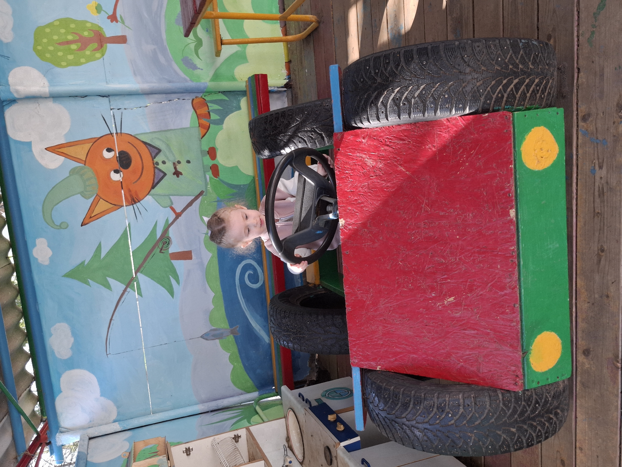 The scene is a playground in the city of Chaikovsky, Russia. A young girl is sitting in a homemade car made of tires and painted wooden planks, holding the steering wheel. The car is red, green, and blue. The girl has blonde hair and wears a pink jacket. Behind the car is a painted wall with a cartoon cat, a tree, and other elements of a landscape. The playground appears to be partly covered, as there are wooden planks above.