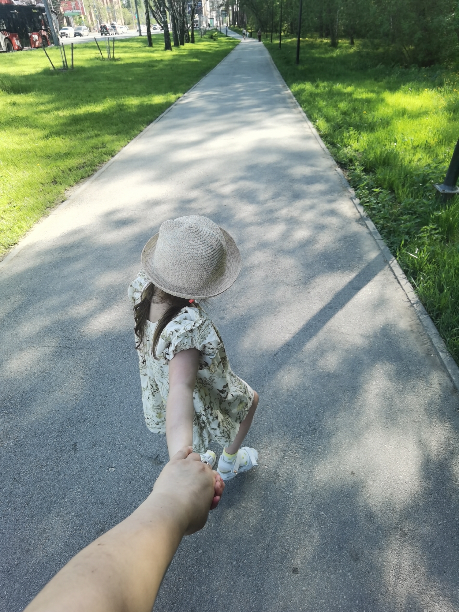 A photograph depicts a first-person perspective of an adult holding the hand of a child on a pathway. The child, wearing a light-colored floral dress and a straw hat, is walking slightly ahead. The pathway is gray and appears to be made of asphalt or concrete. Green grassy areas border both sides of the pathway, lined with trees and occasional street lights. The location is Perm, Russia.