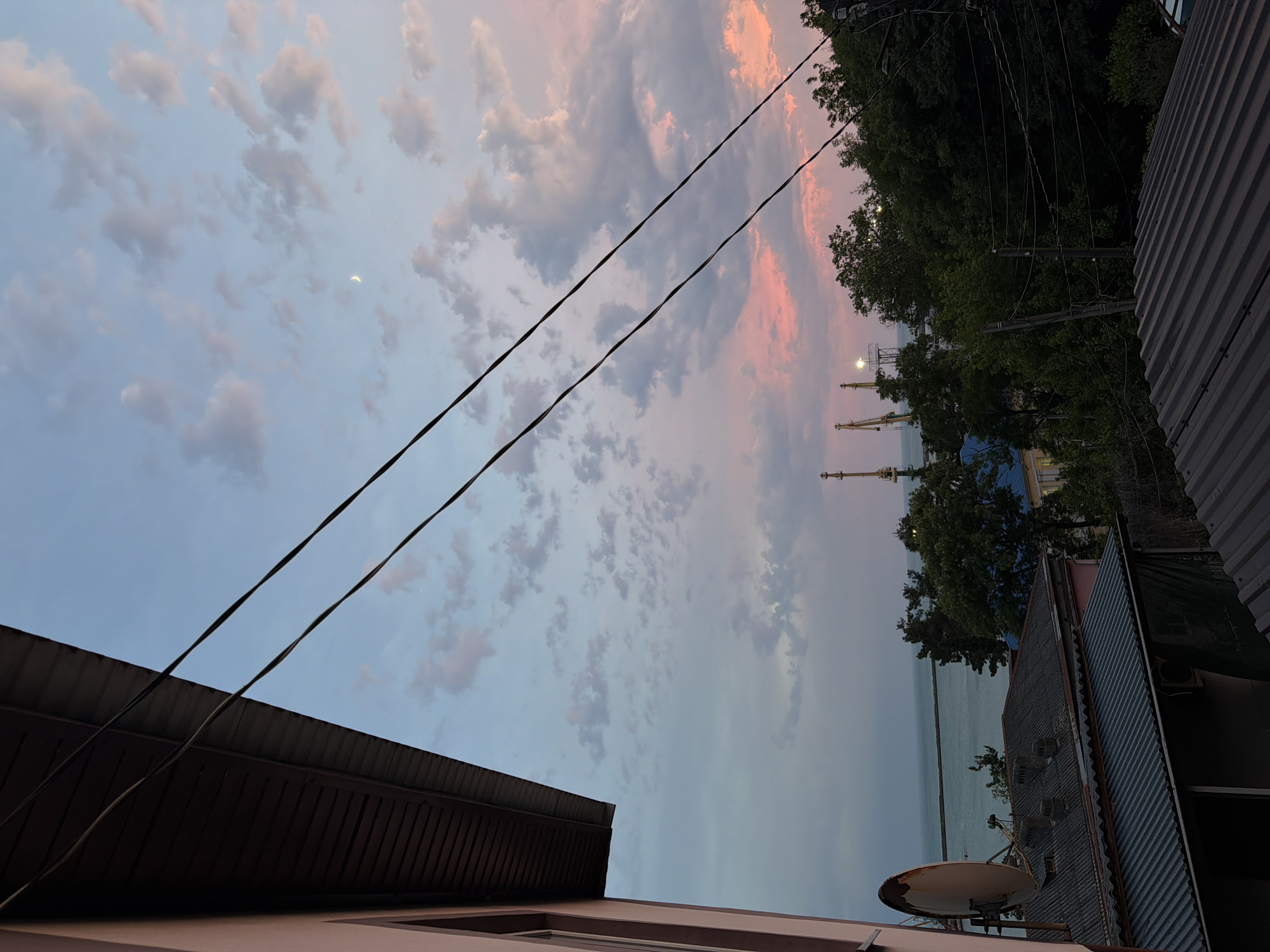 The image captures a dusk scene from a balcony in Tuapse, Russia, overlooking a port and the Black Sea. In the foreground, a pink-colored building's roofline and a satellite dish are visible. Beyond the rooftops, the calm, blue waters of the Black Sea stretch towards the horizon. The middle ground is dominated by dense green trees, behind which rise several yellow cranes, indicative of a port or industrial area. The sky is a gradient of soft blues, pinks, and grays, with scattered clouds and a faint crescent moon visible. Power lines crisscross the sky, adding a utilitarian element to the otherwise serene landscape.