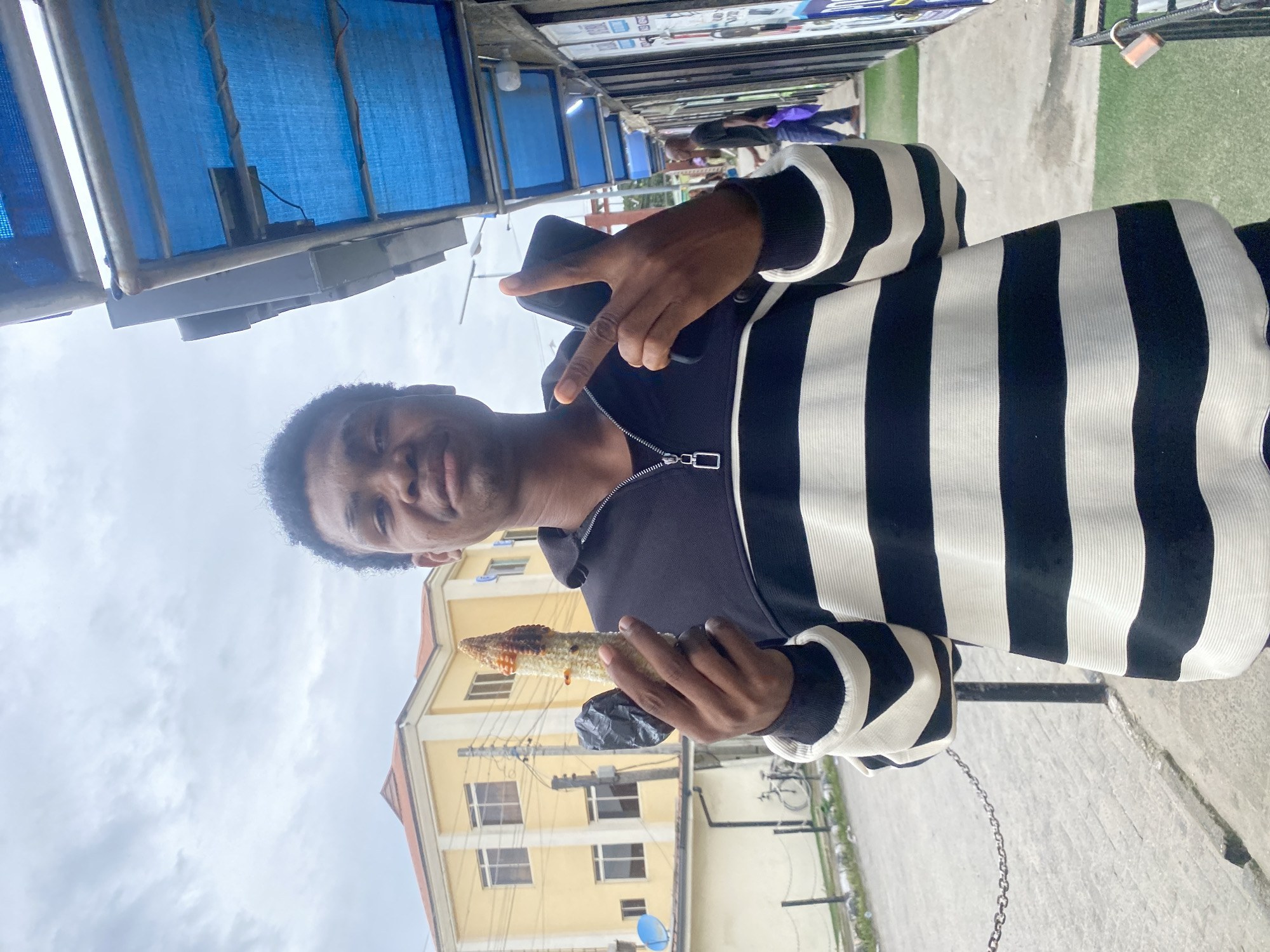 A young Black man stands outdoors in Ado, Nigeria, holding a piece of grilled corn in his left hand and a smartphone in his right. He is wearing a black and white horizontally striped long-sleeved shirt with a zipper at the neck. The man is facing the camera with a neutral expression. Behind him, a yellow building with multiple windows and a red-tiled roof is visible, along with a blue satellite dish. To his right, a row of shops with blue awnings lines the street, and a chain-link fence with a lock is in the foreground. The sky is overcast. The ground is paved with stones and has patches of green artificial turf.