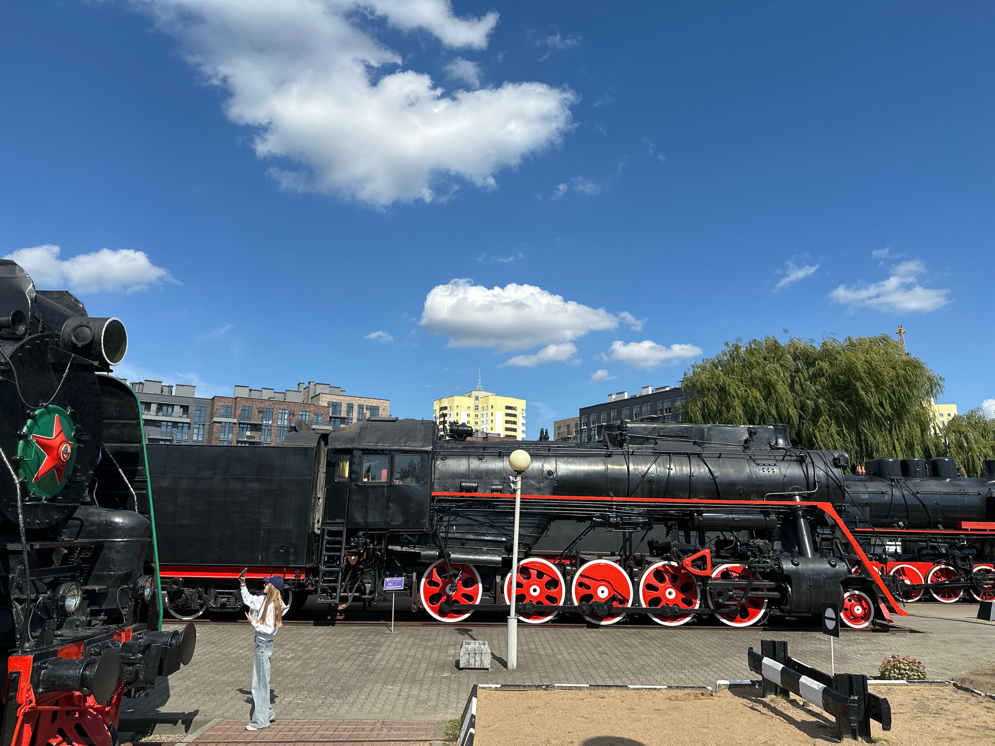 The image depicts an outdoor scene featuring two vintage black steam locomotives parked on a paved walkway under a bright blue sky with scattered white clouds. A young woman wearing a white hoodie, blue jeans, and a baseball cap is standing between the two locomotives, facing the front of the locomotive on the left, with her arm raised as if taking a photo or waving. The locomotive on the left is partially visible, showing its front end with a red and green emblem. The larger locomotive on the right is fully visible, characterized by its prominent red-and-white wheels, a red stripe along its body, and a black chimney. In the background, modern buildings with multiple stories and leafy trees can be seen, suggesting an urban setting. The location is identified as the Museum of Railway Technology in Brest, Belarus. The overall atmosphere is bright and sunny, indicating daytime.