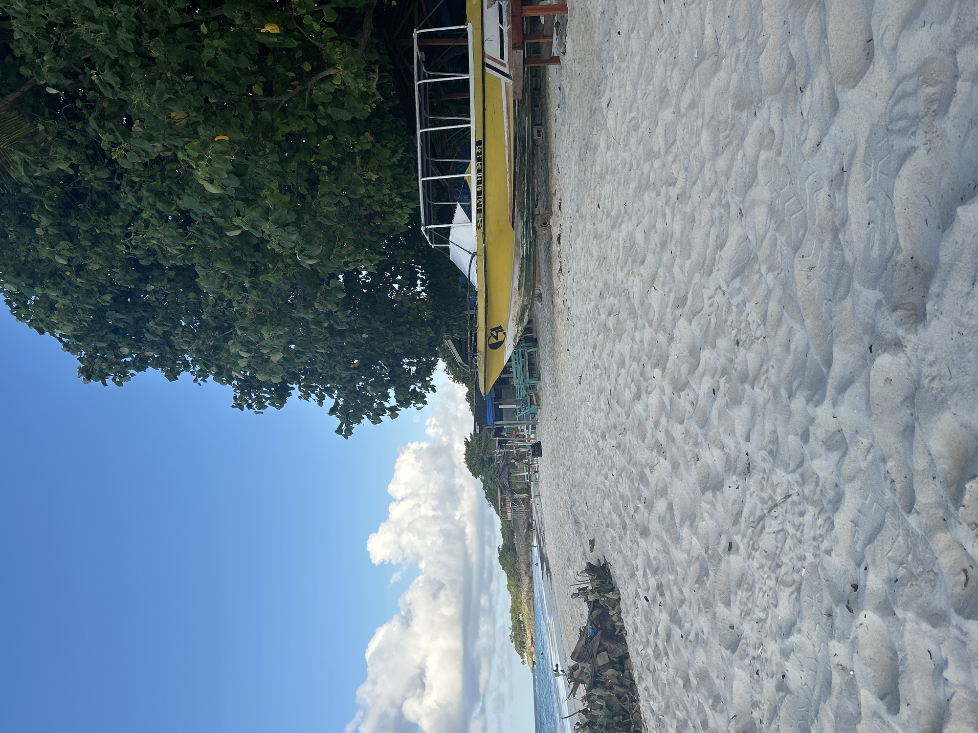The image shows a tropical beach scene. In the foreground, white sand is textured with footprints and scattered debris. A yellow boat with a white canopy sits on the sand, partially covered by a large tree with dense green foliage. In the background, the turquoise ocean meets a coastline dotted with vegetation and several small structures, including thatched-roof huts and a covered seating area. The sky is bright blue with scattered white clouds. The scene appears to be a public beach with recreational facilities. The location is not in A R A, Indonesia, but rather a tropical coastal area.