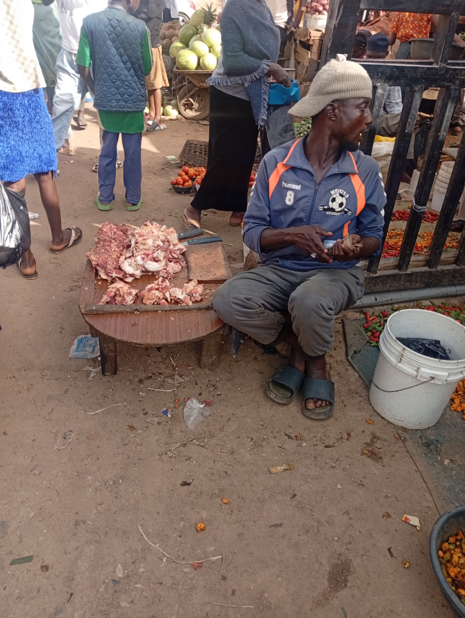 A man wearing a light brown knit cap and a blue and orange jacket sits on the ground beside a low wooden table. He is holding money in his hands. The table is covered with a pile of raw meat, some of which appears to be chopped. To the right of the man is a white plastic bucket, and next to it, a pile of small red and yellow peppers. In the background, a cart filled with large green fruit, possibly watermelons, is visible, along with other market stalls and people. The ground is dirt and littered with debris. This scene is likely from a market in Rafin Par, Nigeria.