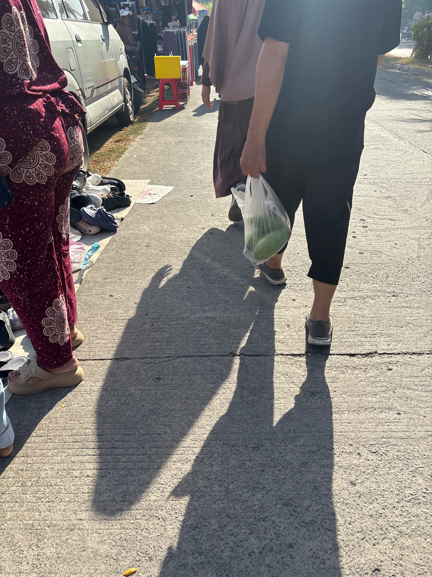 The image depicts a sunny street scene in Telagamurni, Indonesia, where a pedestrian walks alongside a parked van. In the foreground, a person wearing a patterned maroon garment and sandals is partially visible, with a collection of footwear scattered on the ground beside them. Further down the paved street, another individual in dark clothing is seen carrying a clear plastic bag containing two green objects, possibly fruits or vegetables. A third person, in a muted purple garment, walks slightly ahead of the second individual. The background shows a row of stalls with merchandise and trees, suggesting a market or roadside shopping area. The harsh sunlight casts long shadows of the pedestrians onto the concrete surface.