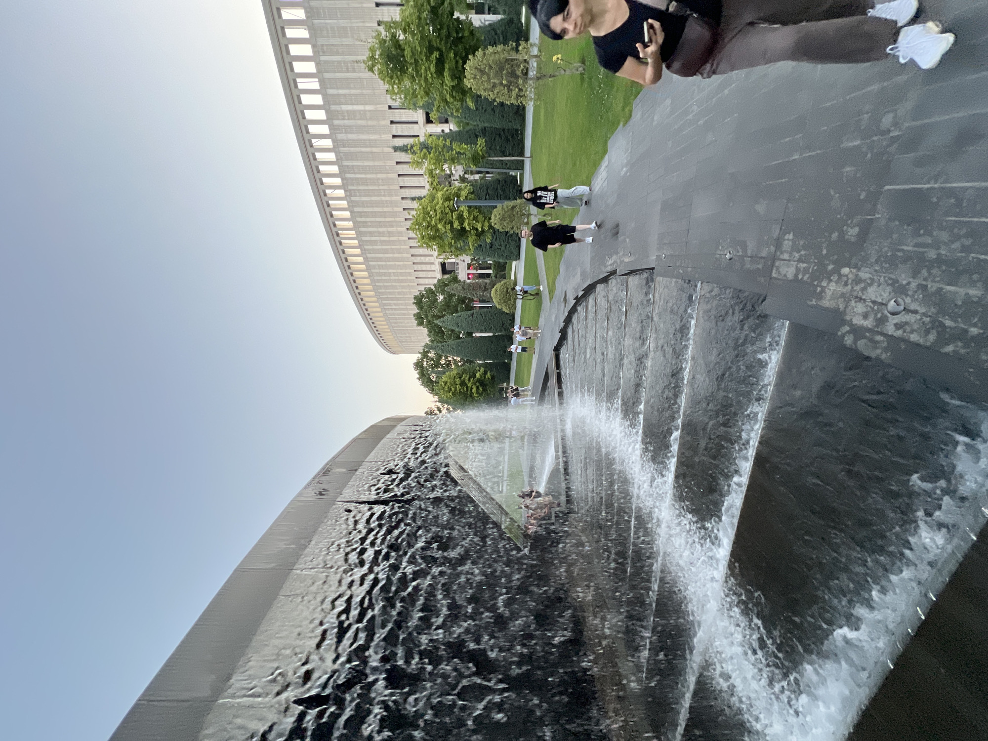 In this photograph, a large, modern building with a rhythmic facade of horizontal lines and recessed rectangular windows dominates the background. The building appears to be an arena or stadium, curved in shape. In front of it, a lush green park with manicured trees and grassy areas suggests a well-maintained public space.
The foreground is occupied by a striking tiered fountain. Water cascades down a series of dark, possibly stone, steps, creating a dynamic and refreshing visual. The water's movement is captured with a sense of motion, with splashes and spray visible.
Several people are present in the scene. In the mid-ground, a group of individuals are seen walking on a paved pathway in the park area. Closer to the fountain, another group is gathered, and one person is partially visible in the upper right corner, looking towards the fountain and holding a phone, possibly taking a picture. The people's attire suggests casual wear, indicating it's a pleasant day for outdoor activities.
The lighting indicates it is daytime, likely late afternoon or early evening, given the soft light and absence of harsh shadows. The sky is clear and a pale blue, suggesting fair weather. The overall atmosphere is one of leisure and public enjoyment of an urban park and its water features. The presence of the grand architectural structure and the energetic fountain points to a location of public significance, possibly a landmark in Krasnodar, Russia. No visible text is present in the image.