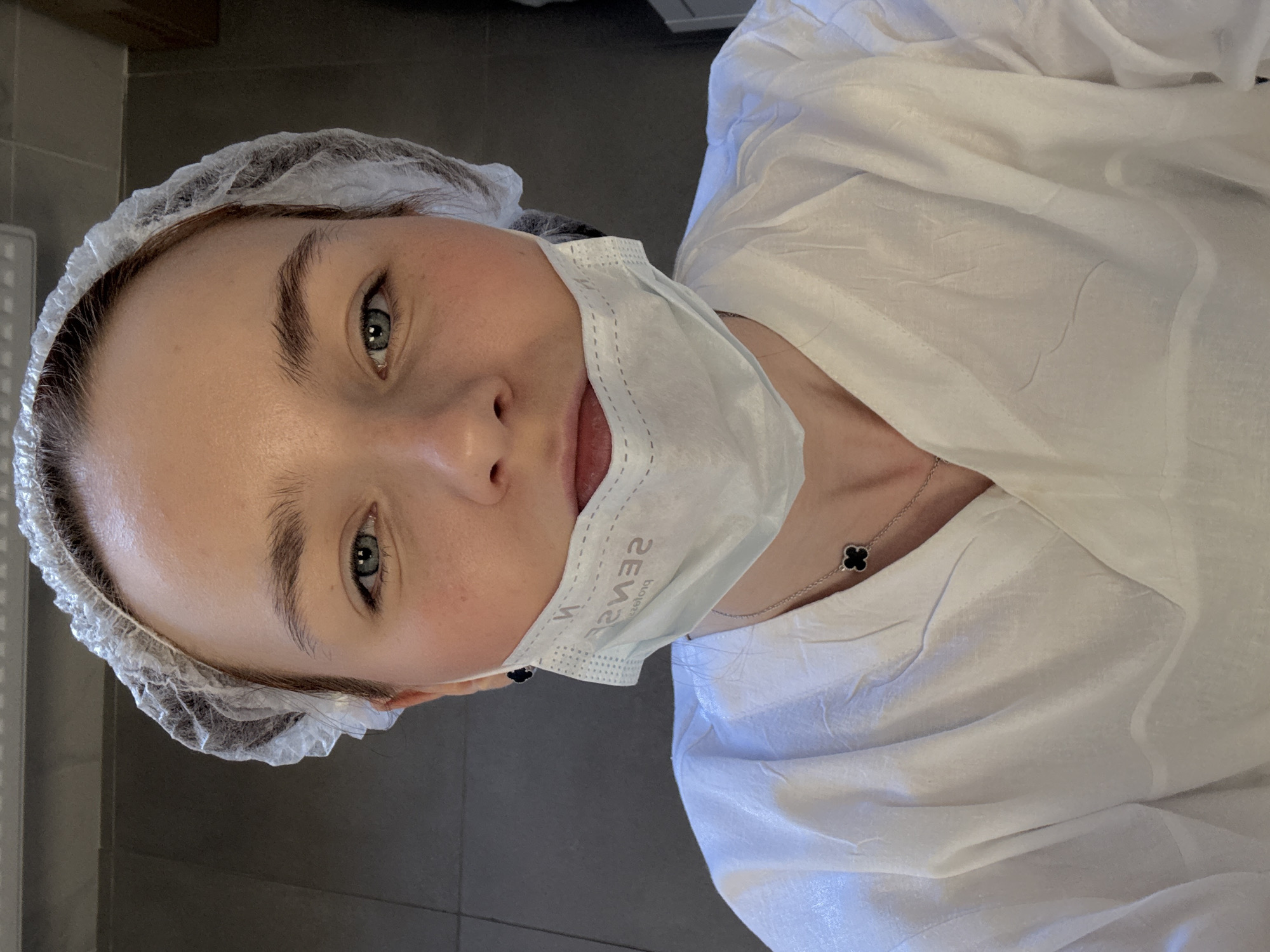 A woman with fair skin, wearing a hairnet and a blue surgical mask labeled "SENSE," looks at the camera. She is also wearing a white garment that may be a lab coat or gown. A necklace with a clover-shaped pendant is visible. The background appears to be a neutral, tiled wall, suggesting an indoor setting, possibly a medical facility or a spa. The image is vertically oriented and the location is unknown but presumed to be in the city of Saint Petersburg, Russia.