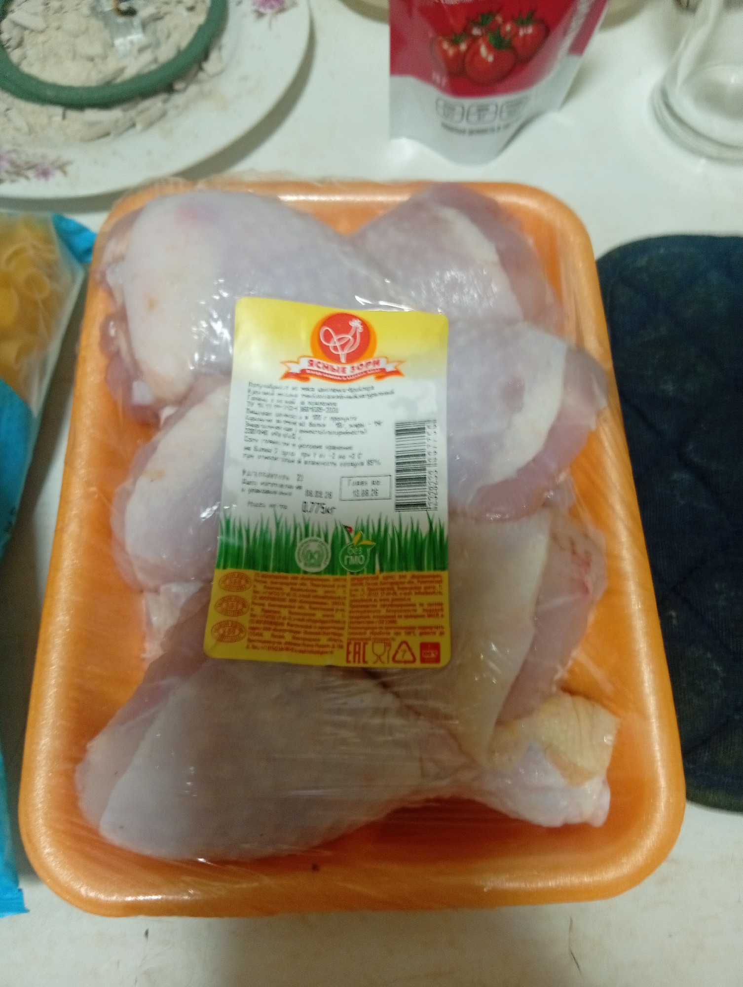 A tray of raw chicken drumsticks is presented in a plastic overwrap. A yellow and orange label is affixed to the plastic, featuring text and imagery including a stylized chicken and a field of grass. The chicken drumsticks are light pink and white, with visible skin and bone. The tray is orange plastic. The background shows a glimpse of a white plate with floral patterns and what appears to be a green coil object. To the left, a portion of a clear plastic bag containing yellow pasta is visible. To the upper right, a red and white package with images of strawberries is partially in view. The scene is set on a white countertop. The overall impression is of a food product in a kitchen or grocery store setting. The specific location in Pushkino, Russia, cannot be determined from the visual information alone.