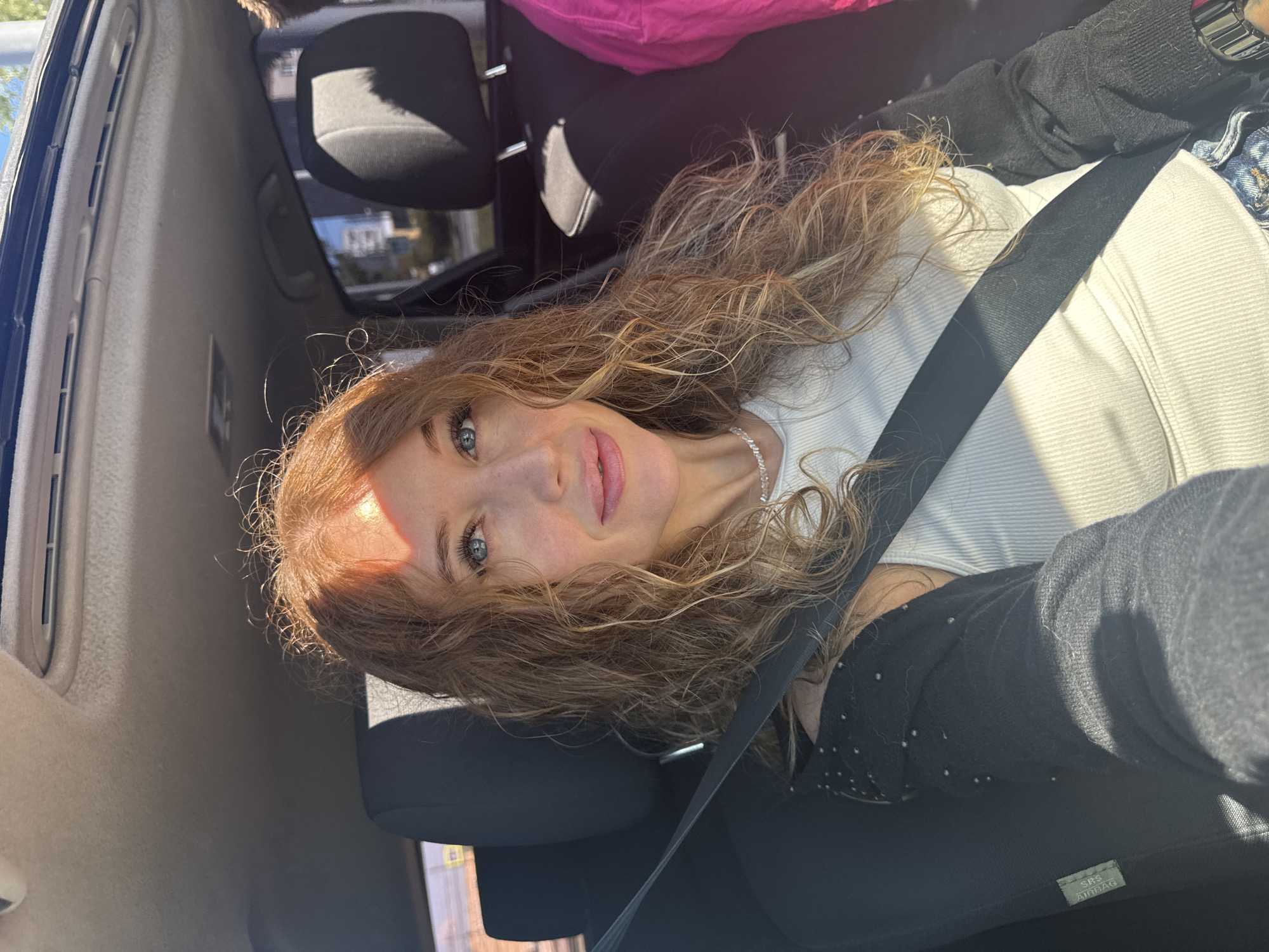 A woman with long, curly auburn hair and blue eyes is sitting in the passenger seat of a car. She is wearing a white ribbed tank top and a black cardigan adorned with small silver studs. A black seatbelt is diagonally across her chest. The interior of the car is visible, including the headrest of the driver's seat and a portion of the car's ceiling. Sunlight streams into the car, casting shadows and illuminating parts of her face and hair. Outside the car, a blurry urban environment is visible through the window, suggesting the car is in motion within the city of Chaikovsky, Russia.