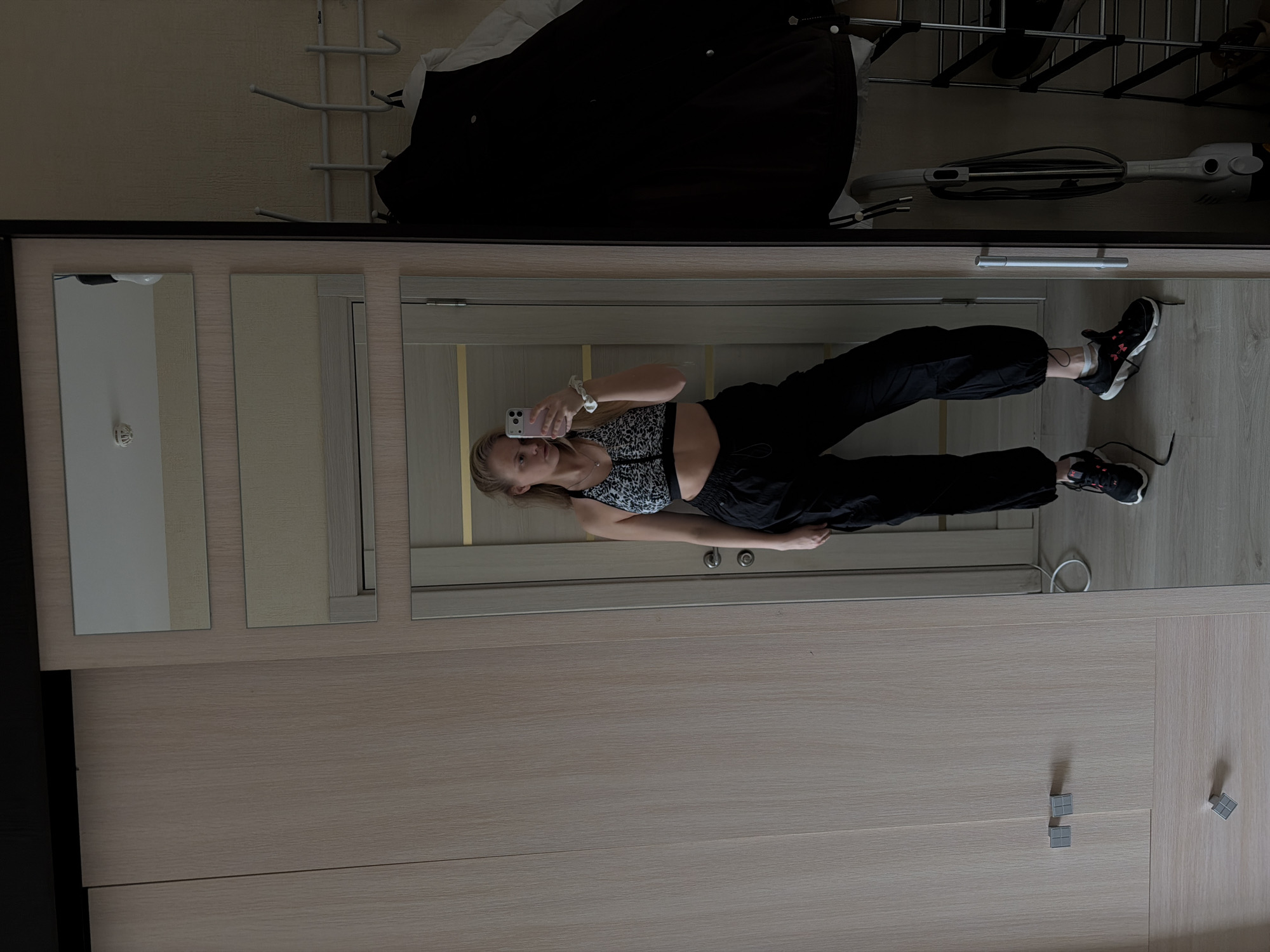 A mirror selfie taken in an interior setting resembling a bedroom in Izhevsk, Russia, shows a young woman dressed in a cropped black and white animal print top and black sweatpants, posing in front of a wardrobe. She holds a smartphone to capture the image. The wardrobe has a mirrored front, reflecting the interior space, including what appears to be a clothing rack with items hanging on it. The room's color scheme includes shades of beige and light brown for the wardrobe and door. The flooring is also a light wood tone. The composition emphasizes the wardrobe and the woman's attire.