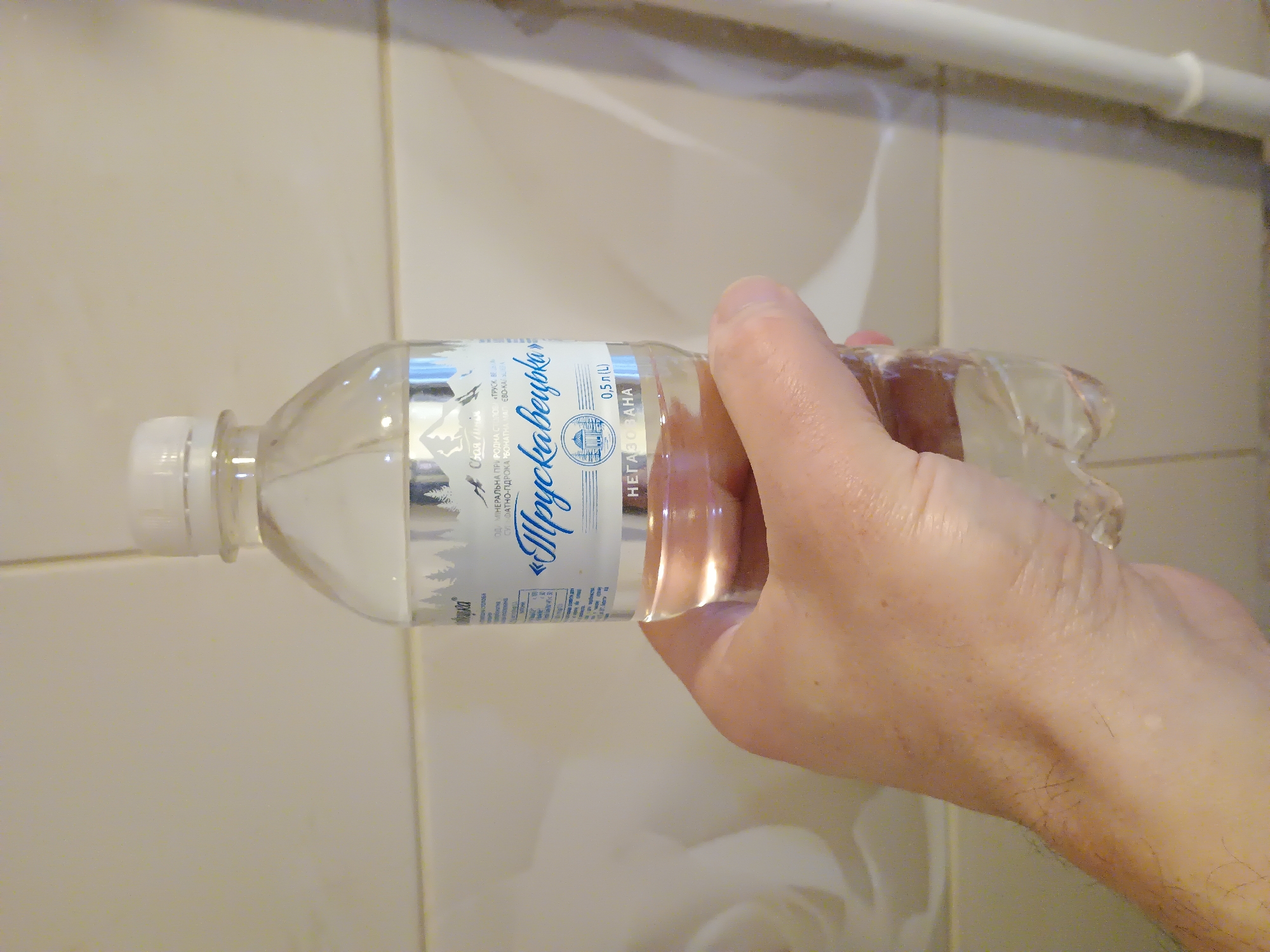 A hand is holding a clear plastic bottle of mineral water with a white cap. The bottle has a label with text in blue and silver. The label features a picture of mountains and trees. Text on the label is in Ukrainian. The bottle contains clear liquid. The background appears to be a tiled wall. The location of Zaporizhzhia, Ukraine, is not depicted in the image.