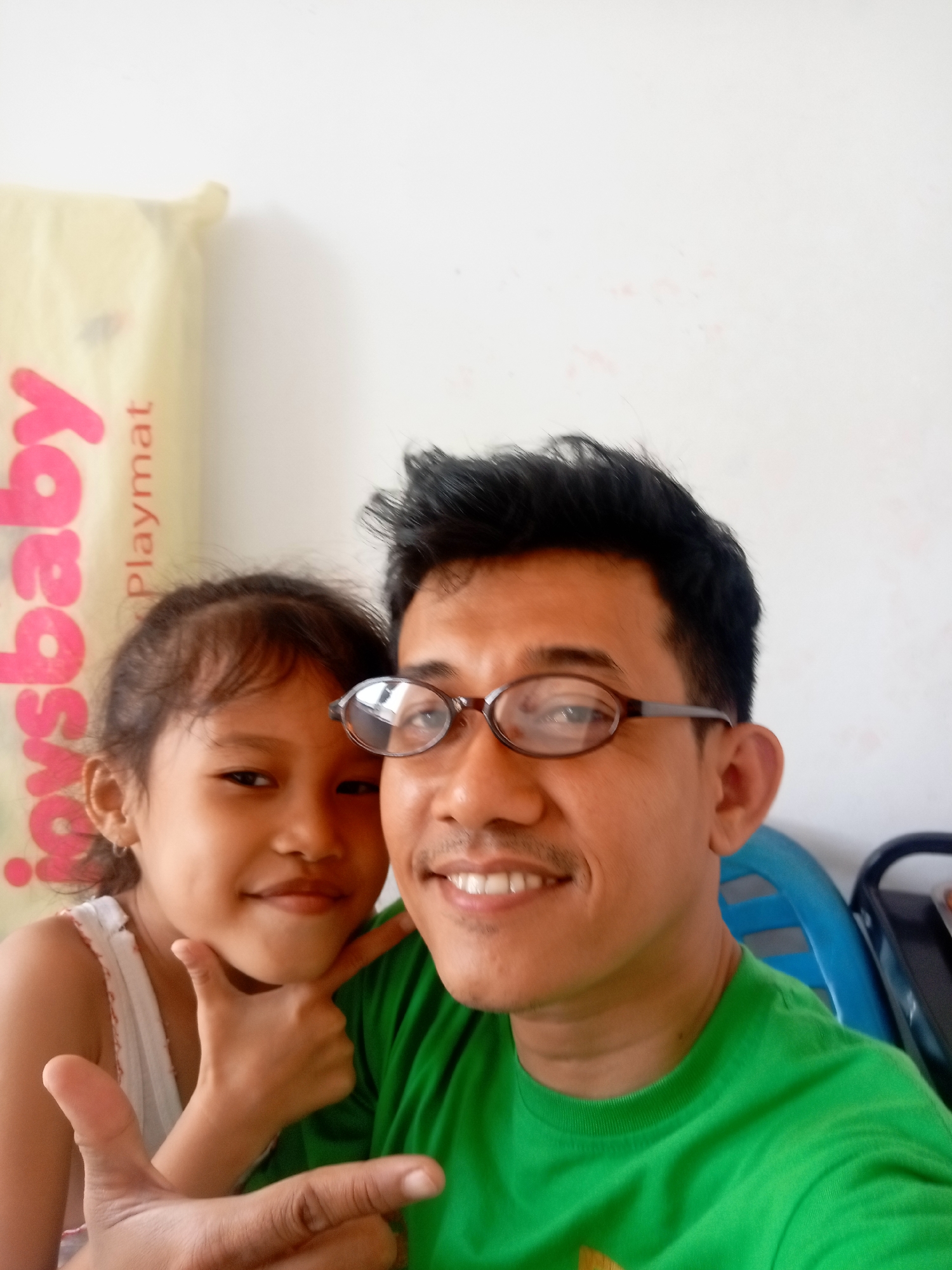 In a close-up selfie, a man wearing a green t-shirt and glasses smiles as a young girl with her hand on her chin poses beside him. The girl is wearing a white tank top. Behind them, a light-colored mat with pink lettering that appears to read "ysbaby Playmat" is visible. The background is a plain white wall with some light red smudges. The setting suggests an indoor environment, possibly a home, in Batam, Indonesia. The man's right hand is extended forward, giving a thumbs-up gesture. The girl's left hand is also positioned near her face, supporting her chin. Both individuals are looking towards the camera.