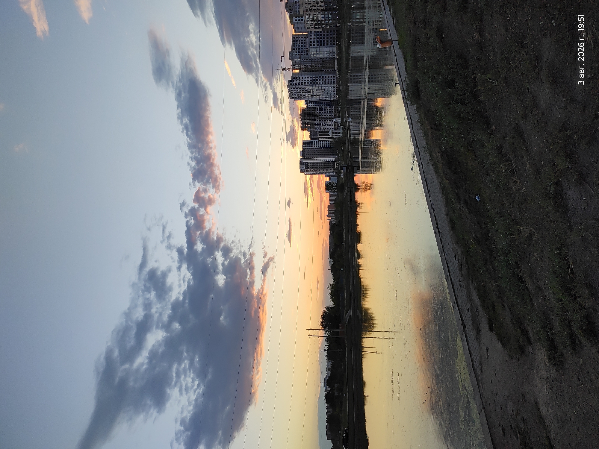 The image shows a waterfront scene at dusk, likely in Astana, Kazakhstan, as indicated by the timestamp "3 авг. 2026 г., 19:51" which translates to "August 3, 2026, 19:51". The sky is a gradient of pale blue and orange, with scattered dark and light clouds. In the distance, a row of modern apartment buildings, some under construction with cranes visible, lines the opposite bank of a body of water. The water reflects the sky and the buildings, creating a mirror-like effect. The foreground consists of a dark, grassy embankment leading down to a paved walkway along the water's edge. There are no people visible in the image, but an orange trash receptacle is present near the right edge of the walkway. The overall atmosphere is calm and serene, characteristic of a city at the end of the day.