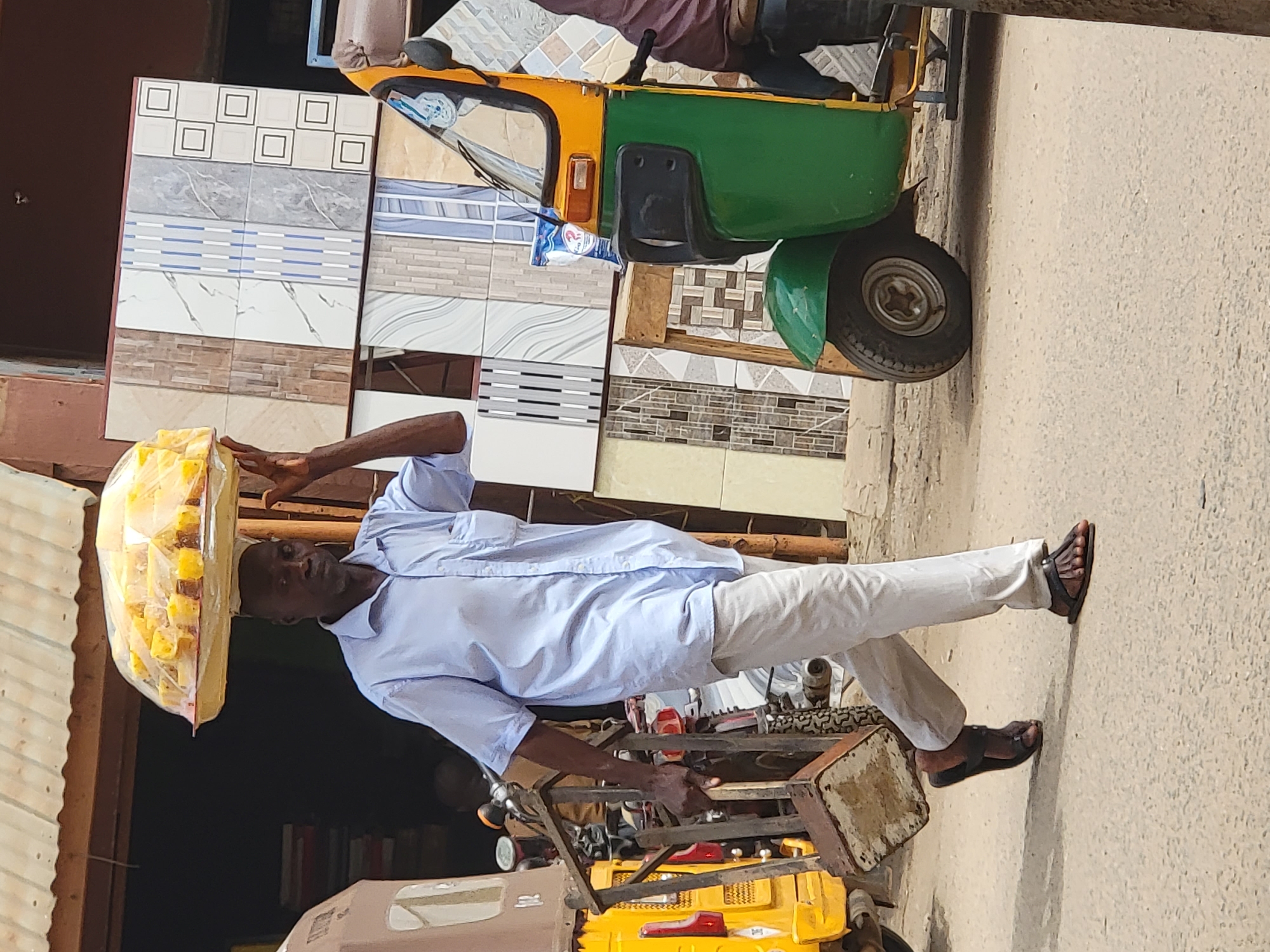 The image, taken in bright daylight in Maiburiji, Nigeria, features a man standing outdoors, skillfully balancing a large, clear plastic bag filled with numerous small, yellow cube-shaped items (likely food products) on his head. He is a dark-skinned man, wearing a light blue long-sleeved shirt, white trousers, and dark sandals. His right hand rests on a metallic frame, which could be part of a cart or another structure.
Behind the man, a yellow and green three-wheeled auto-rickshaw (commonly known as a Keke Napep in Nigeria) is partially visible. A blue and white plastic sachet, bearing the partially visible text "Aji Pure Water," hangs from the auto-rickshaw. In the background, behind the vehicle and the man, there is a prominent display of various patterned and colored wall or floor tiles, suggesting a building material shop or vendor. The ground beneath them is a light, dusty color, and a corrugated metal roof is visible on the far left. The man's neutral expression suggests he is engaged in his daily routine, likely as a street vendor, a common sight in the region.