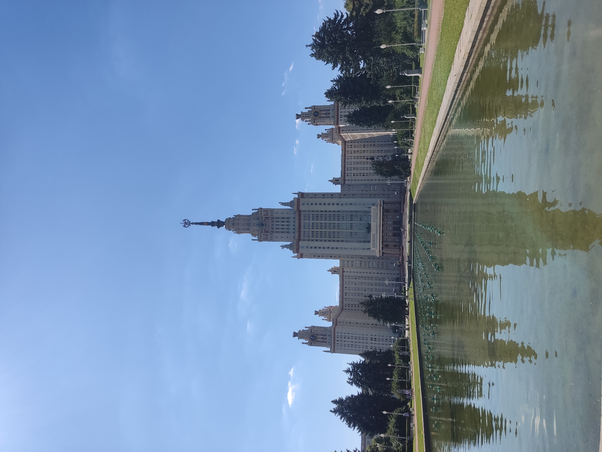 The photograph captures the monumental Stalinist architecture of Moscow State University, located in Moscow, Russia. The main building, a towering edifice with multiple spires and decorative elements, dominates the center of the frame under a clear blue sky with scattered white clouds. In the foreground, a long, rectangular fountain stretches across the width of the image, its calm water reflecting the grandeur of the university building. Rows of small green fountain nozzles are visible in the water. Flanking the fountain are manicured green lawns and pathways lined with lampposts and trees. Several mature coniferous trees are situated between the fountain and the university building. A few indistinct figures of people can be observed in the middle ground walking along a pathway, but they are small and not a focal point of the image. The scene conveys a sense of scale, order, and architectural significance within an urban park setting.
