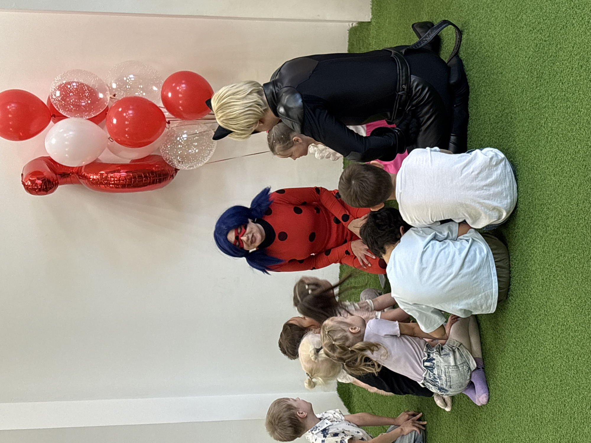 The image shows a group of children gathered on green artificial turf in a room with white walls. Red, white, and clear confetti balloons float near the wall. Two adults dressed as characters, one in a Ladybug costume and the other in a Cat Noir costume, are interacting with the children. The costumes are red with black spots and black with a tail, respectively. The children appear to be engaged with the costumed characters. The environment suggests an indoor play area or party room. The location, Krasnoyarsk, Russia, is not discernible from the image.