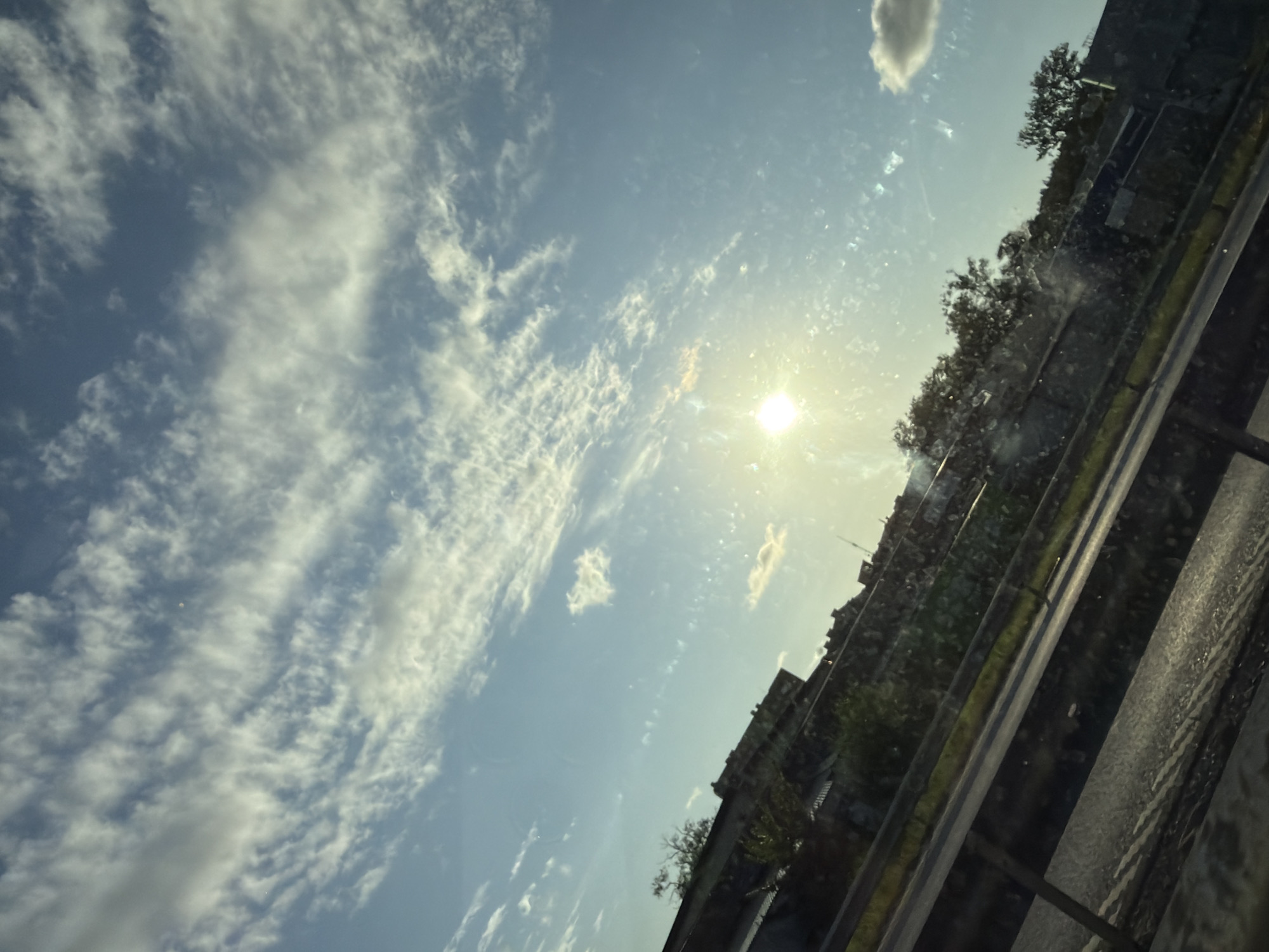 This image shows a view of the sky and a roadside from a vehicle. The sky is blue with scattered white clouds, and the sun is visible and bright. Below the sky, there is a roadside with a guardrail. Beyond the guardrail, there are buildings, trees, and vegetation. The scene appears to be a sunny day, possibly during daylight hours. The perspective suggests the photo was taken from a moving vehicle on a road, given the diagonal lines of the road and guardrail. The presence of buildings and trees suggests an urban or suburban environment. It is not possible to determine the specific location in Mölndal, Sweden from the image alone, as there are no identifying landmarks or signage.
