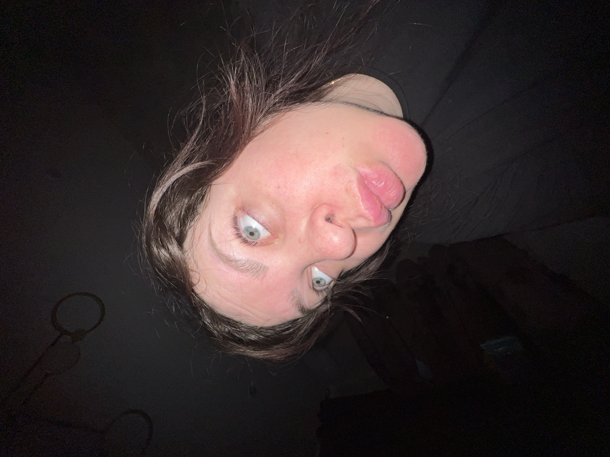 The image is a close-up, low-angle shot of a person's face. The person has fair skin, light-colored eyes, and dark hair. They are making a funny face with their eyes wide and lips pursed. The background is dark and out of focus, with some indistinct shapes that could be objects in a room. The person is wearing a dark-colored shirt. There are no visible landmarks or other details to identify the location as Noviy, Russia, or any specific city.