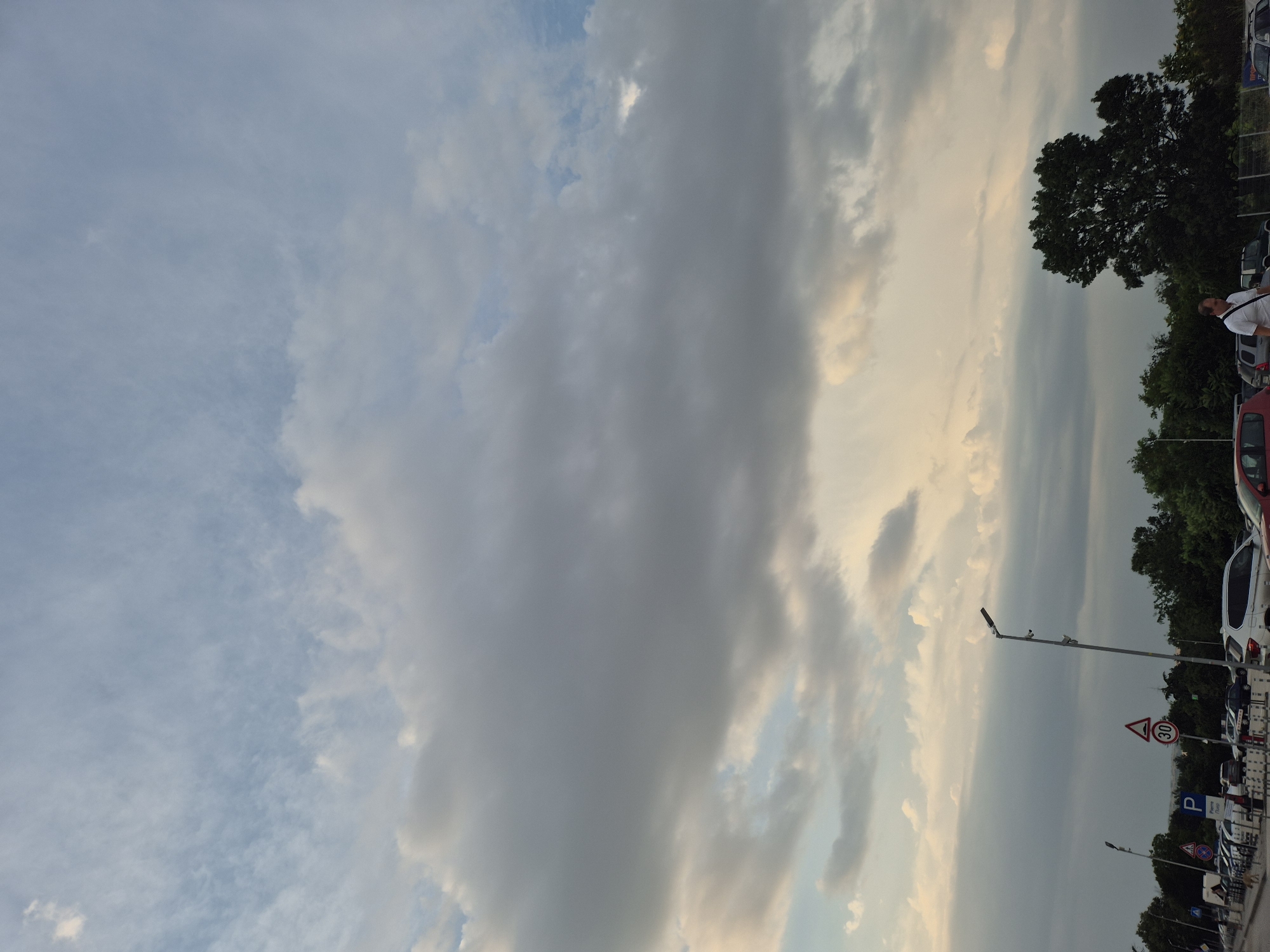 This is a vertical, medium shot of a cloudy sky over a parking lot. The sky is filled with large, grey clouds with some areas of lighter clouds and hints of blue sky peeking through. Towards the right side of the image, there are trees and a line of parked cars. A man in a white shirt is visible on the right side of the frame, facing away from the camera. There are several traffic signs visible on poles, including a sign with "30" inside a red circle and a blue parking sign. The lighting suggests it is late afternoon or early evening, with the sun low on the horizon causing a warm glow on some of the clouds. The overall atmosphere is calm and overcast.