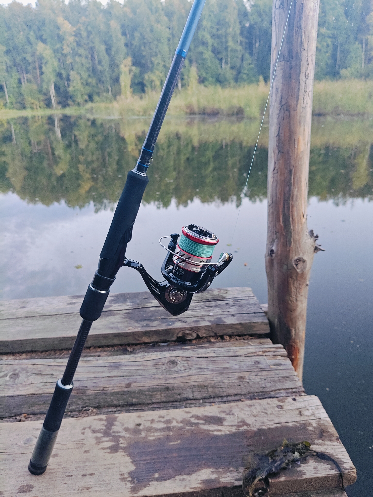 An outdoor, eye-level, medium shot shows a fishing rod and reel resting on a wooden dock next to a body of water. The fishing rod is black and blue, with a black grip and a silver ring. The reel is black and silver, with a spool of light green fishing line. The dock is made of weathered wooden planks. Behind the dock, a calm body of water reflects the trees and sky. The trees are green and lush, and the sky is overcast. In the background, a rough-hewn wooden post stands vertically. The scene appears to be a serene fishing spot, possibly in Chaikovsky, Russia, given the context.