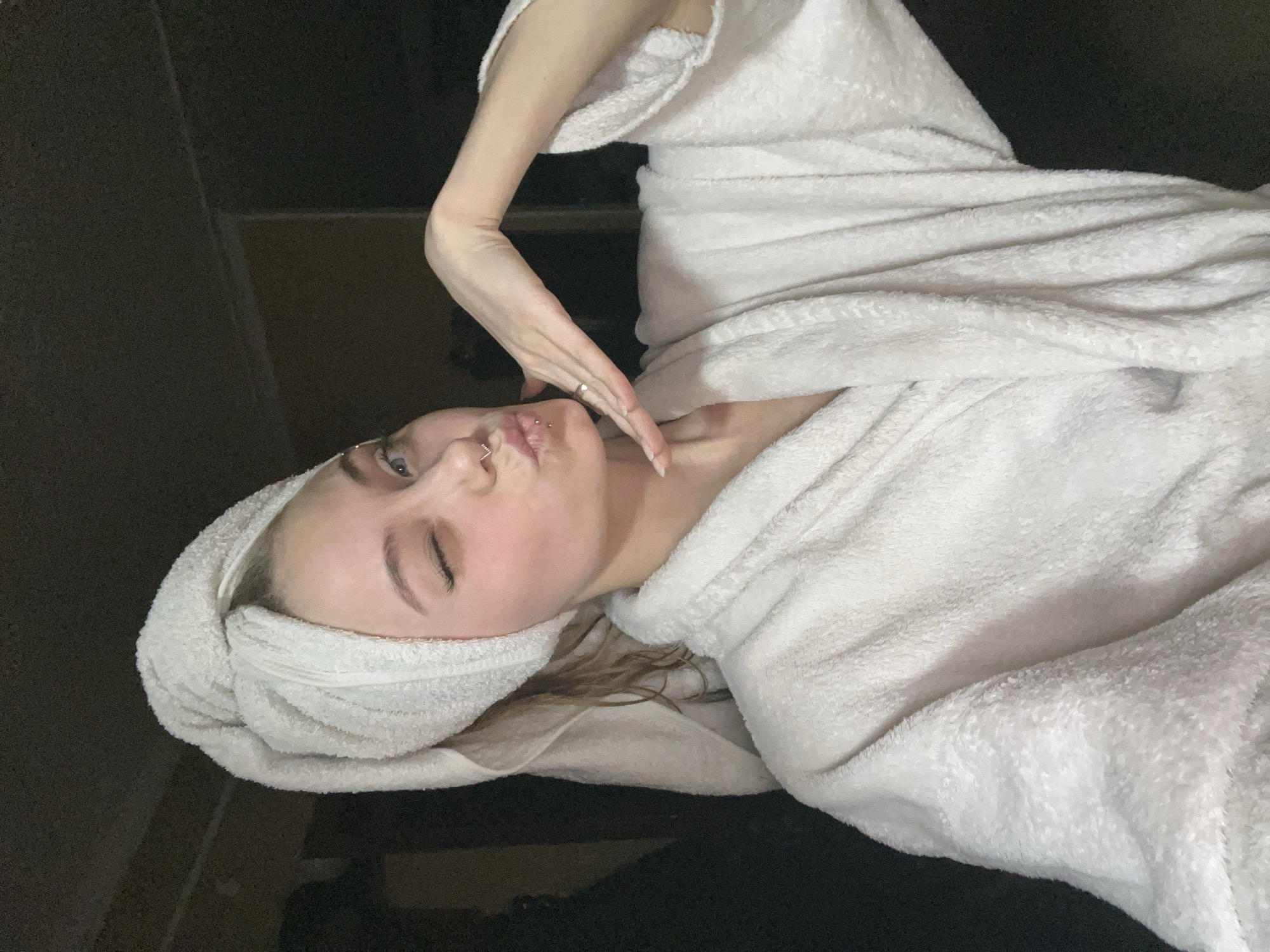 A young woman is lying down, wrapped in a white towel, with another towel wrapped around her head. She has piercings on her nose and lip, and is touching her face with her hand. The background appears to be a dark room. I cannot verify this scene is set in the city of Фоки, Russia.