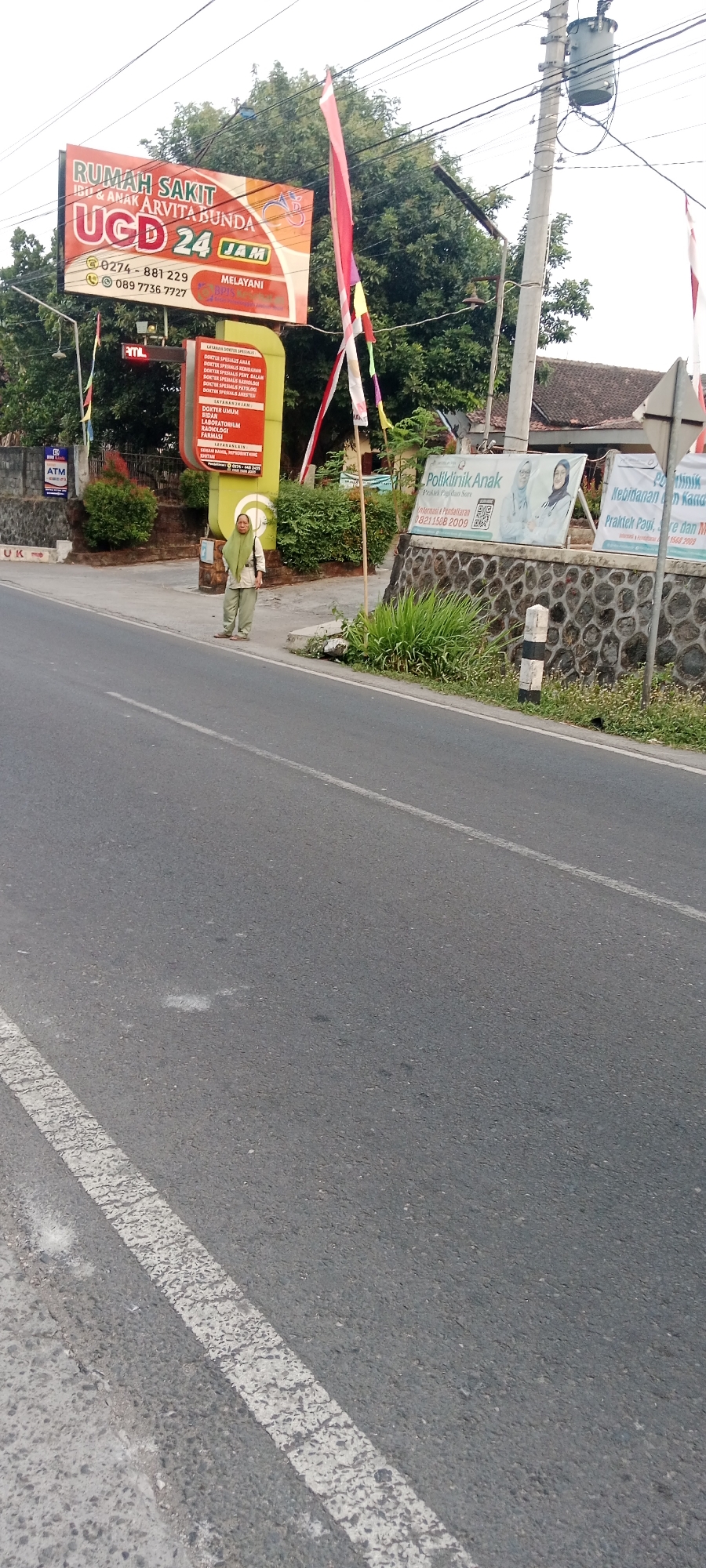 In Maguwoharjo, Indonesia, a street scene unfolds with a prominent "RUMAH SAKIT IBU & ANAK ARVITA BUNDA" sign, indicating a hospital with a 24-hour UGD (Emergency Room) service. Adjacent to this, a yellow sign details various medical services and specialist doctors. To the right, a "Poliklinik Anak" (Pediatric Clinic) sign displays an advertisement featuring two women, one in a white hijab and the other in a blue hijab, with a QR code and contact information.
A woman wearing a light green headscarf and matching loose clothing stands on the sidewalk, facing slightly away from the camera, with her hands clasped in front of her. She appears to be observing the surroundings. The environment includes a paved road with white lane markings, a stone wall, and lush green foliage. Overhead, electrical wires and a transformer are visible against a bright sky. Indonesian flags, red and white, flutter from poles near the signs, adding a festive or patriotic touch. The overall impression is one of a typical roadside setting in a developed area, with medical facilities and pedestrian activity.