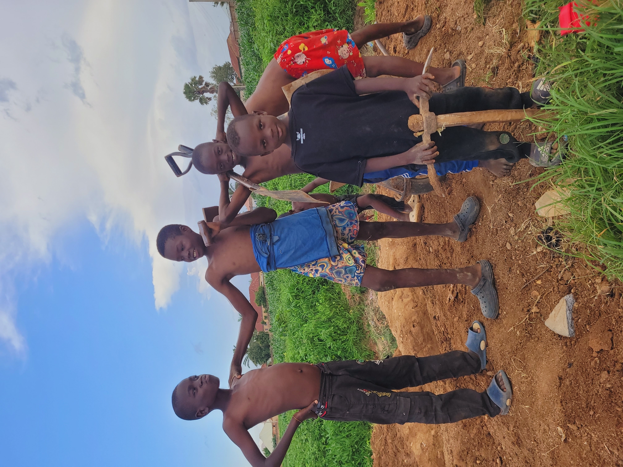 In Bukuru, Nigeria, five young boys are gathered on a mound of brown dirt, possibly engaged in construction or agricultural work. The sky above is a vibrant blue with scattered white clouds. The boys, shirtless and dressed in various shorts and pants, are holding tools that appear to be shovels and picks, suggesting they are working. One boy in the foreground wears a black t-shirt and black track pants with camouflage-patterned sandals, holding a pickaxe. Behind him, another boy in red shorts and dark sandals stands. To his left, two boys stand together, one with a blue cloth wrapped around his waist and patterned shorts, holding a shovel. Another boy to his left, wearing black pants and blue slides, poses with hands on his hips. The environment is green and overgrown with vegetation in the background, with some buildings and a palm tree visible in the distance. The ground is uneven and dusty, indicating a work in progress.