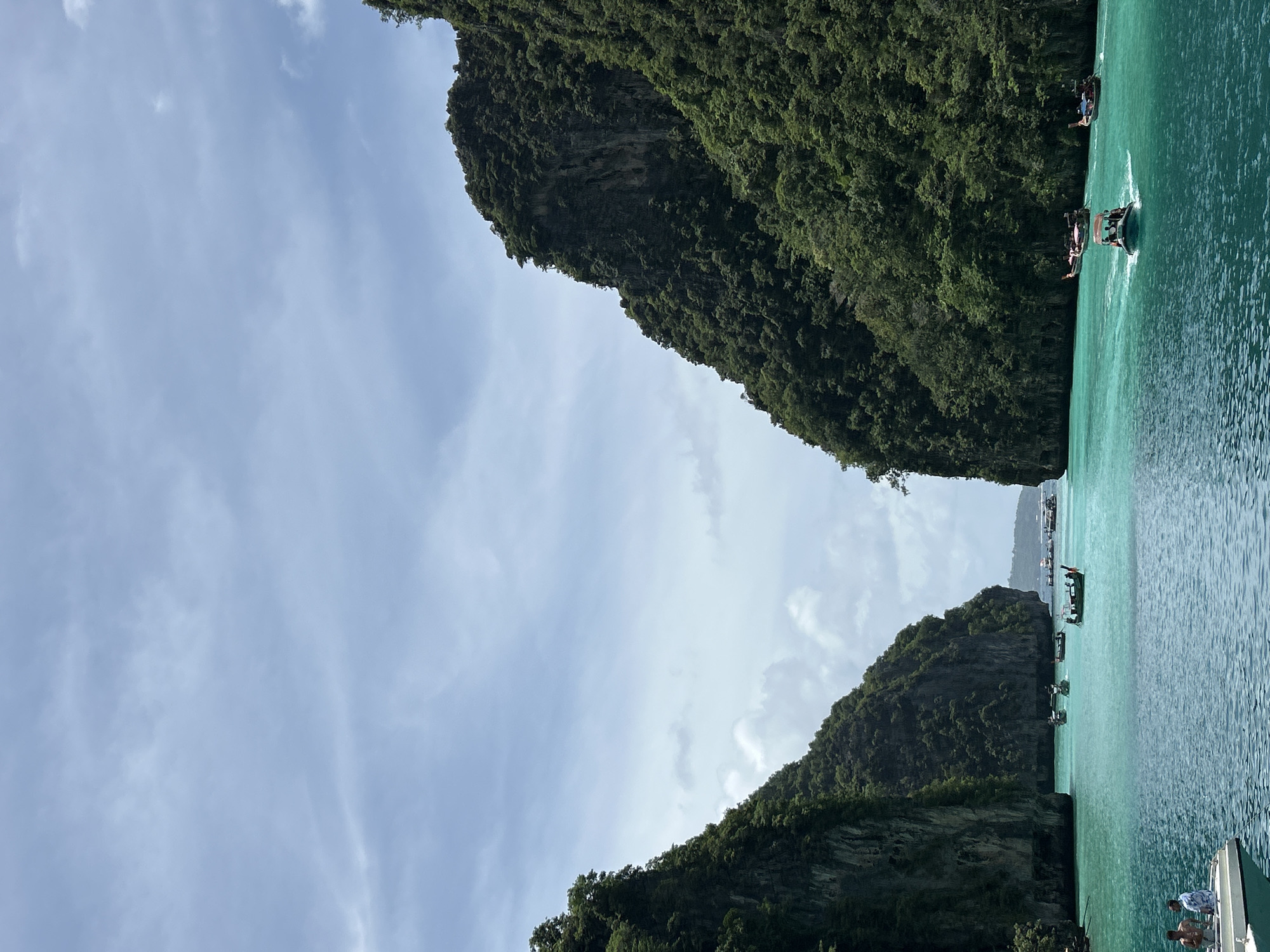This is a vertical, eye-level, full shot of a tranquil bay in Ao Nang, Thailand. The bay is framed by two large, verdant, rocky cliffs that recede into the distance. The water is a clear, turquoise hue, with small ripples on the surface. Several longtail boats, a common mode of transport in Thailand, are scattered across the water. One boat, in the foreground on the left, carries two passengers. Another, in the middle ground, has four occupants, and further back, several more longtail boats are visible, some carrying passengers. The sky is a pale blue, with scattered white clouds. The overall scene is peaceful and picturesque, showcasing the natural beauty of the Thai coast.