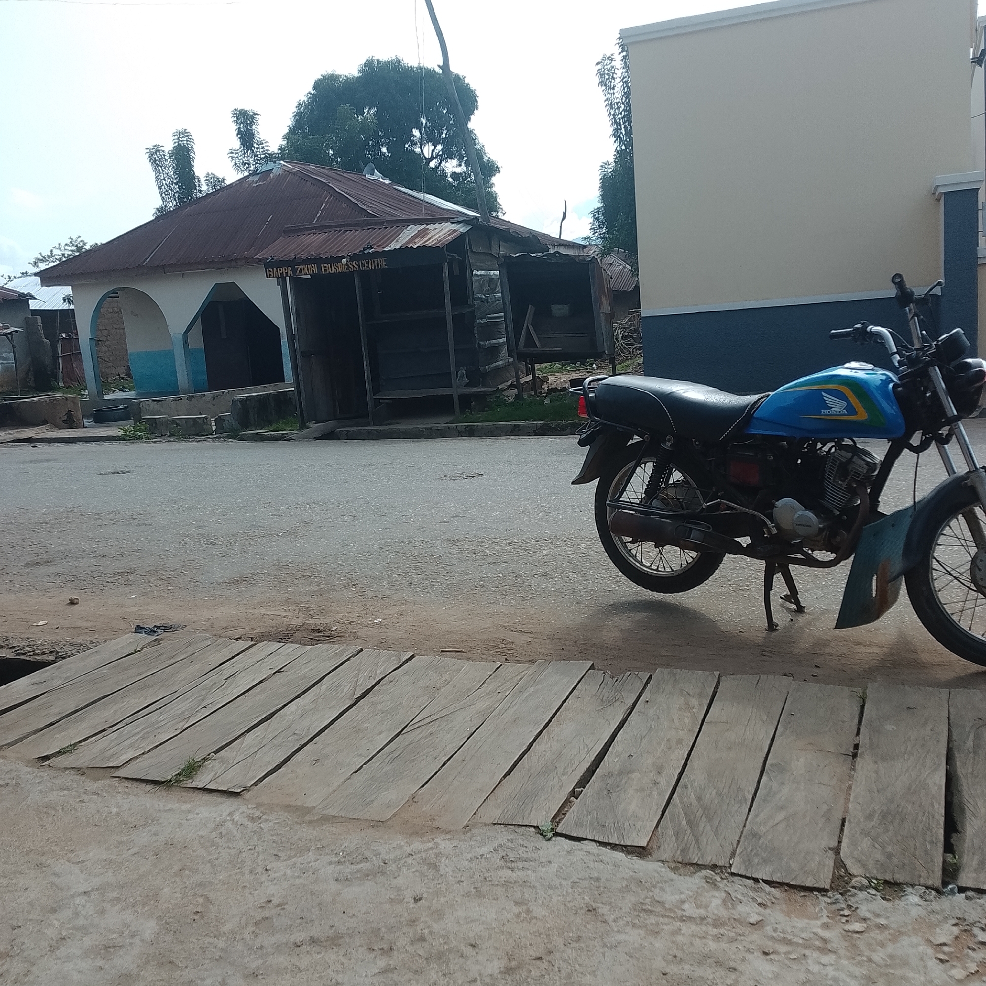 The image shows a street scene in an urban environment. In the foreground, a wooden walkway or bridge spans across what appears to be a ditch or low-lying area. The walkway is made of rough-hewn wooden planks laid side by side. Beyond the walkway, a paved road is visible, with a motorcycle parked on the right side of the frame. The motorcycle is blue and silver with a Honda logo on the fuel tank. In the background, there is a building with a corrugated metal roof, painted white and blue, and next to it, a more modern building with a light-colored facade and a dark blue lower section. Trees and vegetation are visible in the background, suggesting a potentially tropical or sub-tropical climate. The overall impression is one of a semi-developed area.