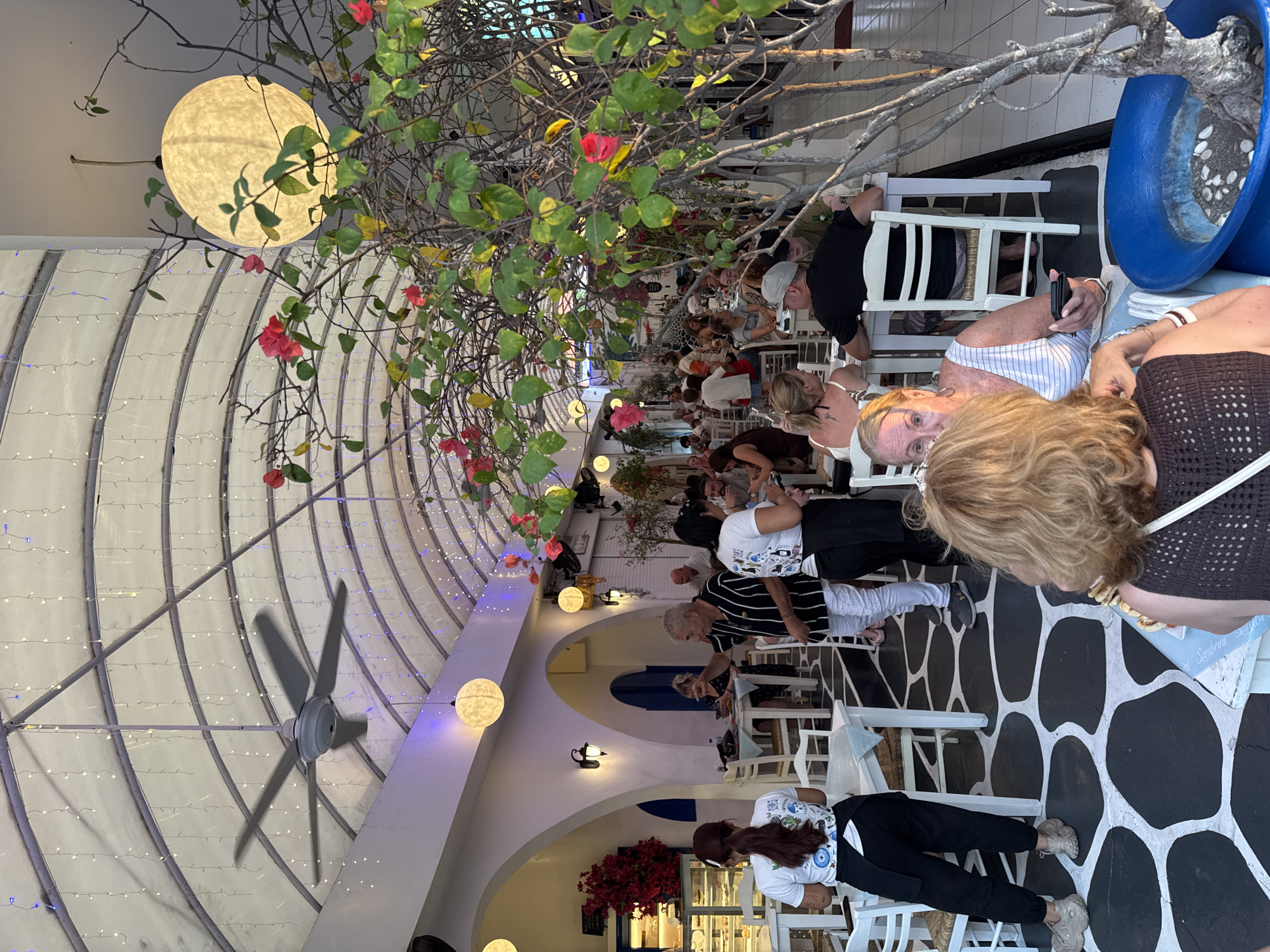 The image depicts a bustling outdoor dining area in Kuta, Indonesia. The scene is vibrant with people seated at tables, enjoying meals and conversation. The architecture features white arched doorways and a white translucent roof supported by curved metal beams, adorned with strings of fairy lights. A large ceiling fan is visible, and several spherical, illuminated lanterns hang from the ceiling. Lush green foliage with bright pink and red flowers drapes over parts of the dining area, adding a natural element. The floor is paved with a distinctive black and white pattern resembling large, irregular stones. Several staff members, identified by their uniforms, are seen serving customers and attending to tables. The overall atmosphere is lively and social, suggesting a popular restaurant or cafe.