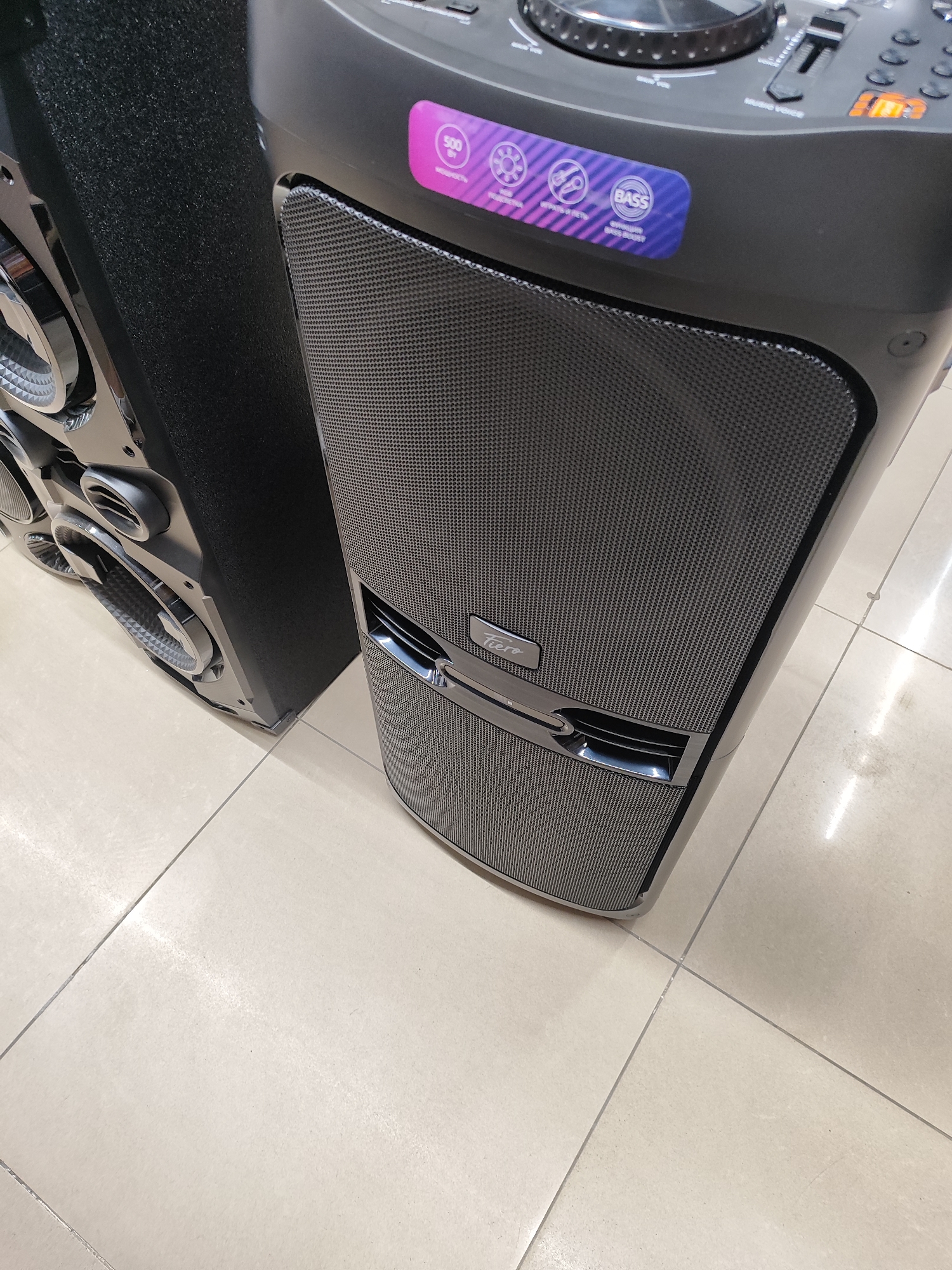 The image shows a close-up view of two large black speakers on a light-colored tiled floor. The speaker on the left is partially visible, showing silver-colored speaker grilles and metallic accents. The speaker on the right is the main subject, displaying a textured black speaker grille on its front. Above the grille is a control panel with a digital display showing "FTO", a slider, and several buttons. A purple sticker with icons and text, including "500", "BASS", and "BASS BOOST", is affixed to the upper part of the speaker. A small metallic emblem with the word "Feero" is centered on the speaker grille. The lighting is bright, casting some reflections on the glossy surfaces of the speakers. The environment appears to be an indoor space, possibly a store or showroom, given the tiled floor and the product display. There are no people visible in the image. The scene is static and focused on the two electronic audio devices.