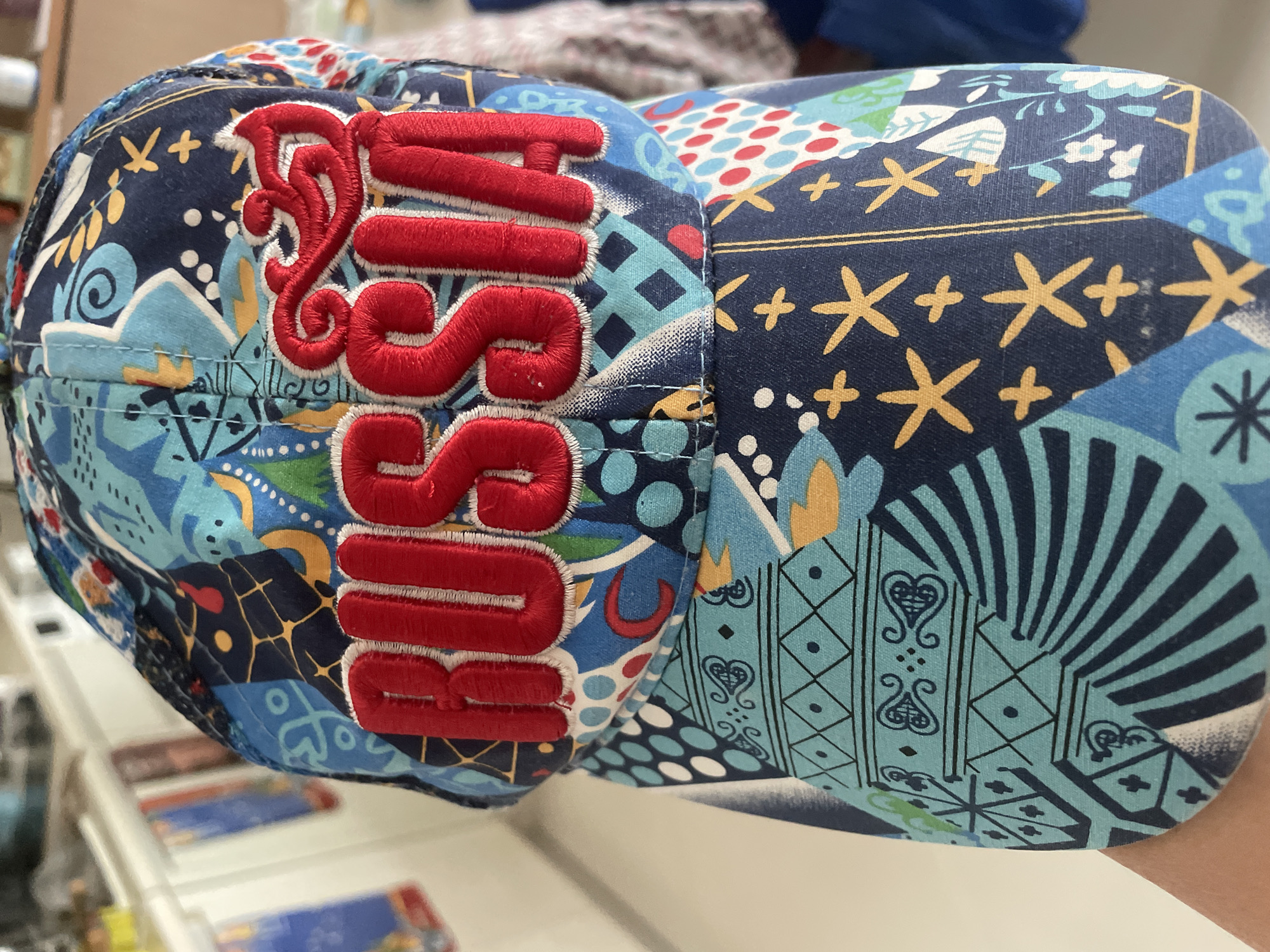 A close-up, eye-level shot shows a colorful baseball cap with the word "RUSSIA" embroidered in red letters. The cap features a busy pattern of abstract shapes, dots, and star-like figures in shades of blue, red, white, and yellow. The background is blurred and appears to be an indoor setting with white cabinetry and other objects, possibly a kitchen or retail space. The image was taken in Pattaya City, Thailand.